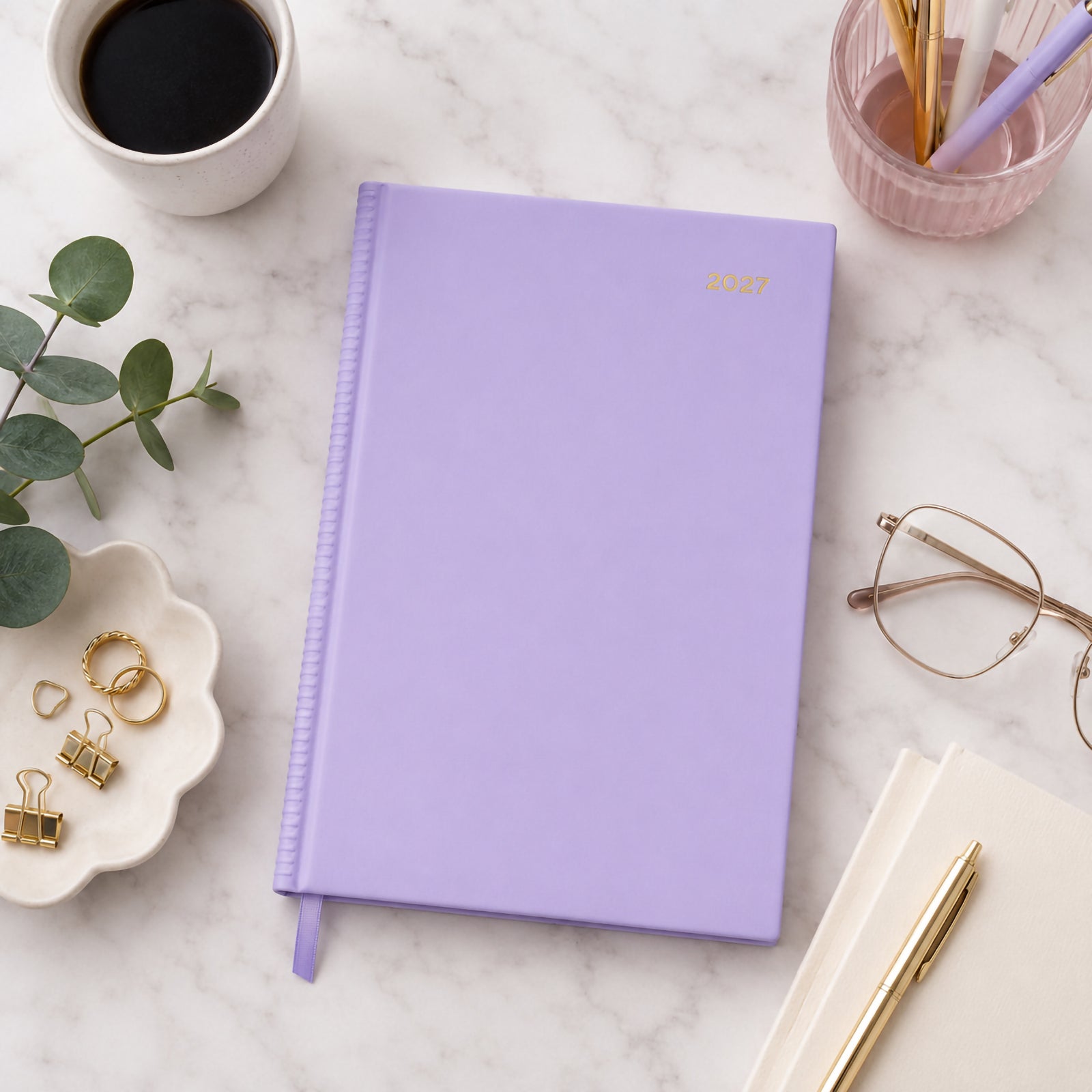 Top view of the Collins Belmont Colours 2027 A5 Day to a Page Lilac Diary resting on a marble desktop with a coffee cup, gold pen, glasses, stationery accessories and eucalyptus leaves. The soft lilac cover creates a fresh and modern planning workspace.