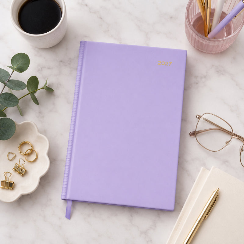 Top view of the Collins Belmont Colours 2027 A5 Day to a Page Lilac Diary resting on a marble desktop with a coffee cup, gold pen, glasses, stationery accessories and eucalyptus leaves. The soft lilac cover creates a fresh and modern planning workspace.