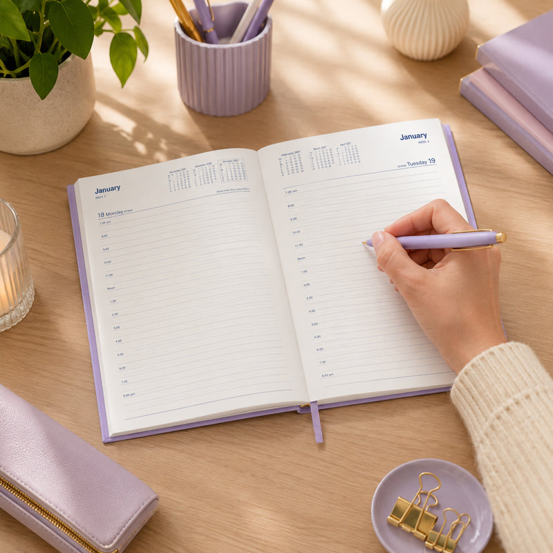 A hand writes inside the Collins Belmont Colours 2027 A5 Day to a Page Lilac Diary on a bright timber desk surrounded by coordinated lilac stationery, plant, organiser accessories and elegant desk decor. The image highlights the spacious daily layout and stylish everyday planning environment.