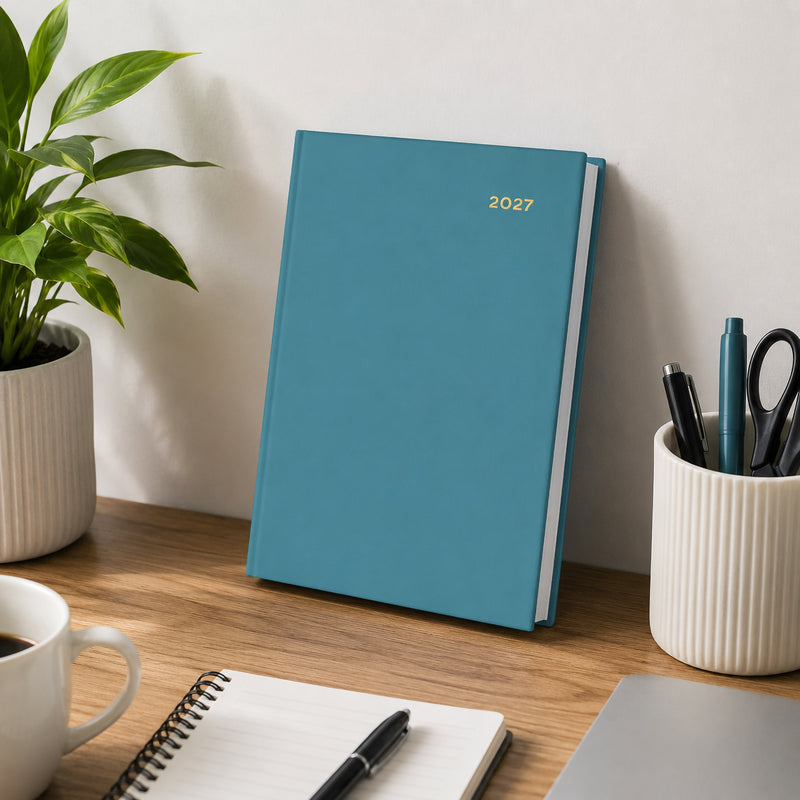 The Collins Belmont Colours 2027 A5 Day to a Page Teal Diary stands upright against a wall on a modern office desk surrounded by stationery, coffee and greenery. The image highlights the elegant teal cover in a professional workspace.