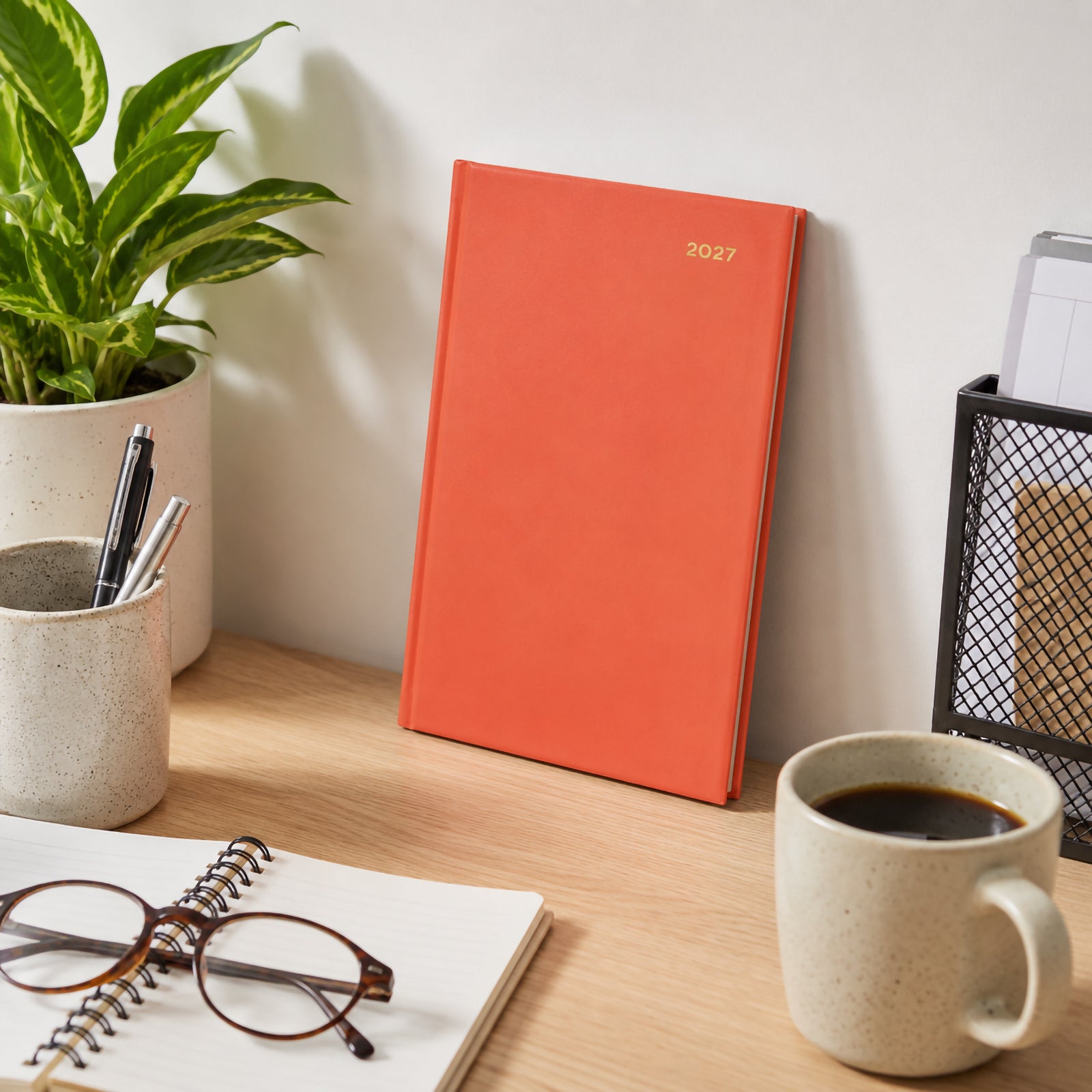 The Collins Belmont Colours 2027 A5 Week to View Orange Diary stands upright against a wall on a tidy desk surrounded by office accessories, a coffee cup, indoor plant and document organiser, highlighting its bright professional appearance.