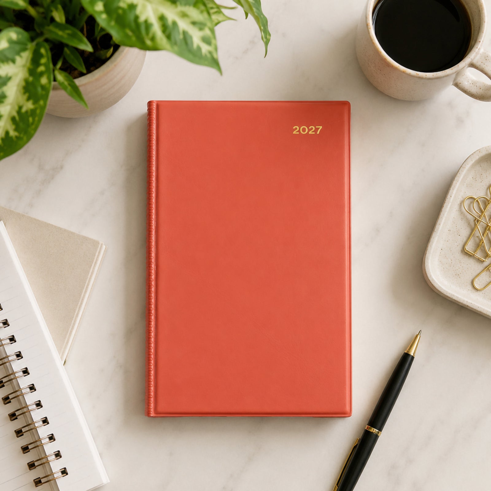 Top view of the Collins Belmont Colours 2027 A5 Week to View Orange Diary positioned on a marble desktop beside a coffee cup, notebook, stationery and indoor plants. The vibrant orange cover adds colour and organisation to a modern office setting.