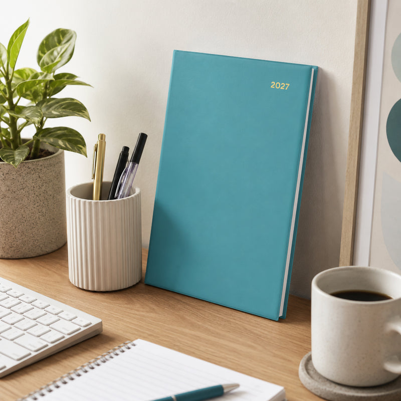 The Collins Belmont Colours 2027 A5 Week to View Teal Diary leans against a wall on a tidy office desk beside stationery, a keyboard, coffee mug and indoor plant, showcasing its elegant teal cover in a contemporary workspace.