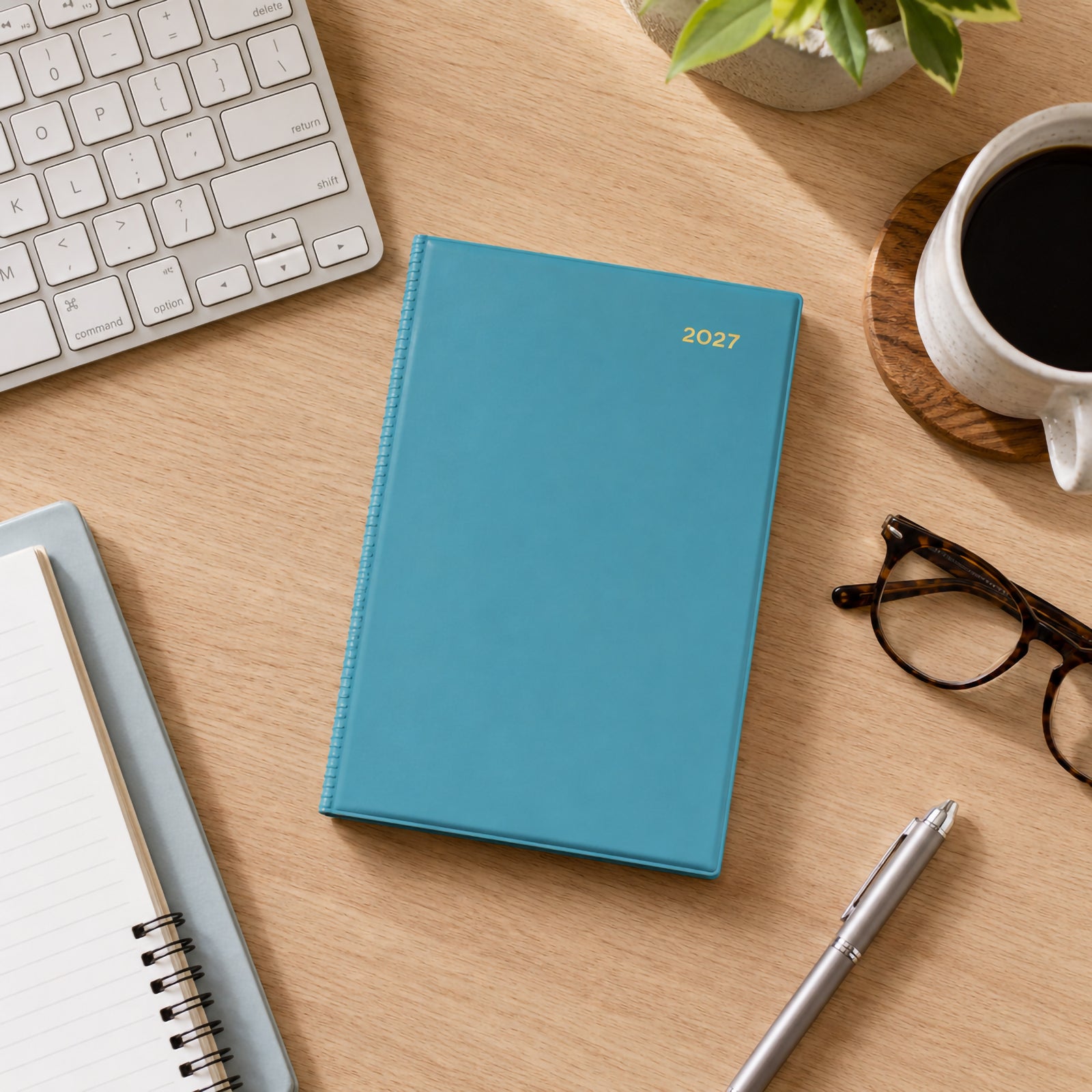 Top view of the Collins Belmont Colours 2027 A5 Week to View Teal Diary resting on a timber desktop with a keyboard, notebook, coffee cup, pen and indoor plant. The calming teal cover complements a clean and organised office environment.