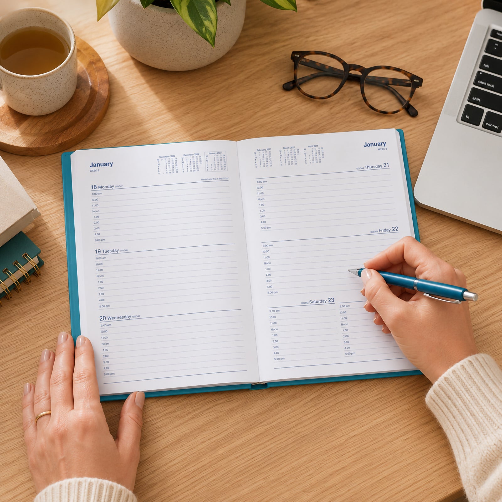 A user writes in the Collins Belmont Colours 2027 A5 Week to View Teal Diary on a modern timber desk. The open weekly planner is surrounded by a laptop, coffee cup, indoor plant and glasses, demonstrating productive daily planning in a professional workspace.