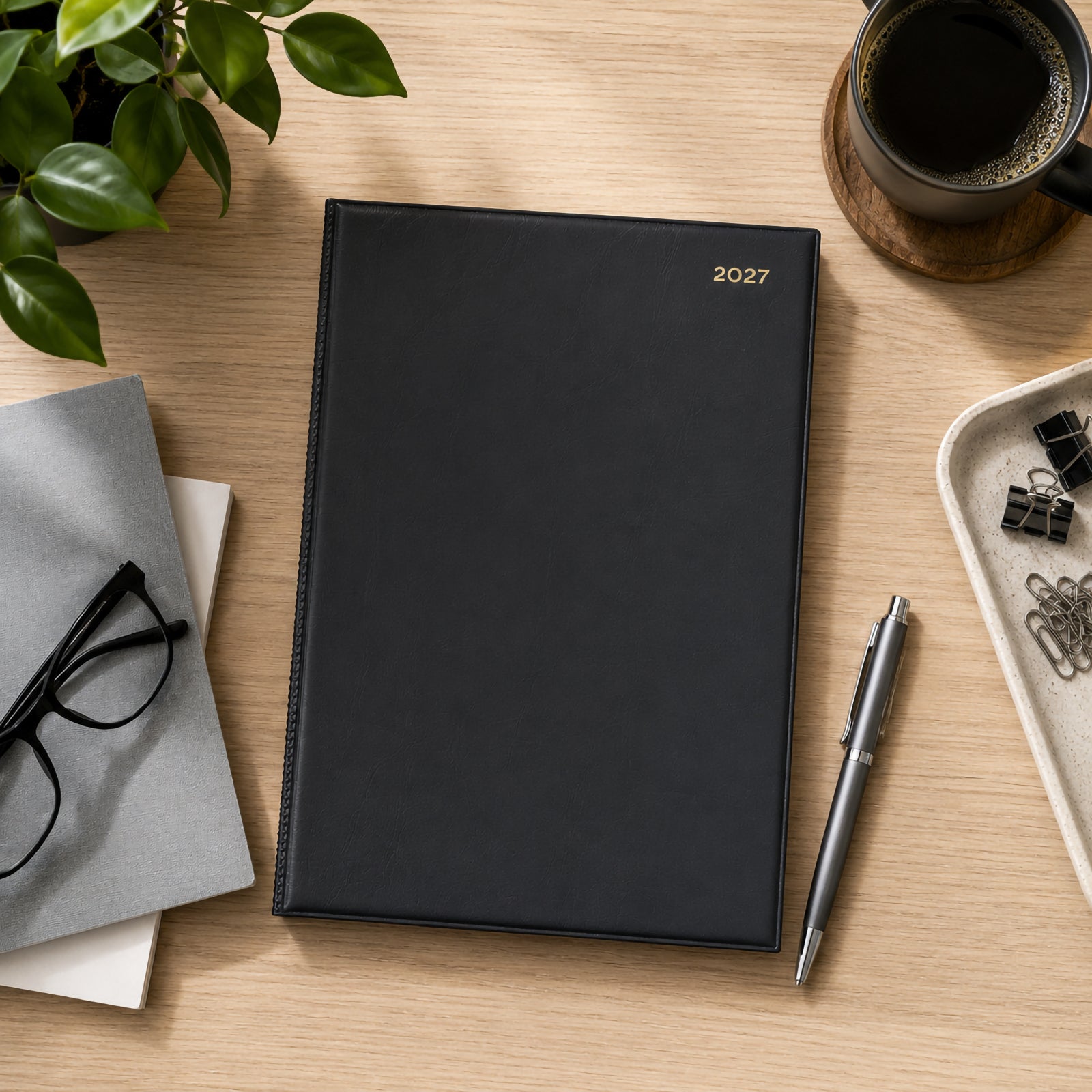 Top view of the Collins Belmont 2027 A4 diary with its black padded PVC cover placed neatly on a timber desk. Surrounded by office stationery, glasses, paper clips, a pen, coffee and a green plant, the diary presents a clean executive appearance suitable for professional planning.