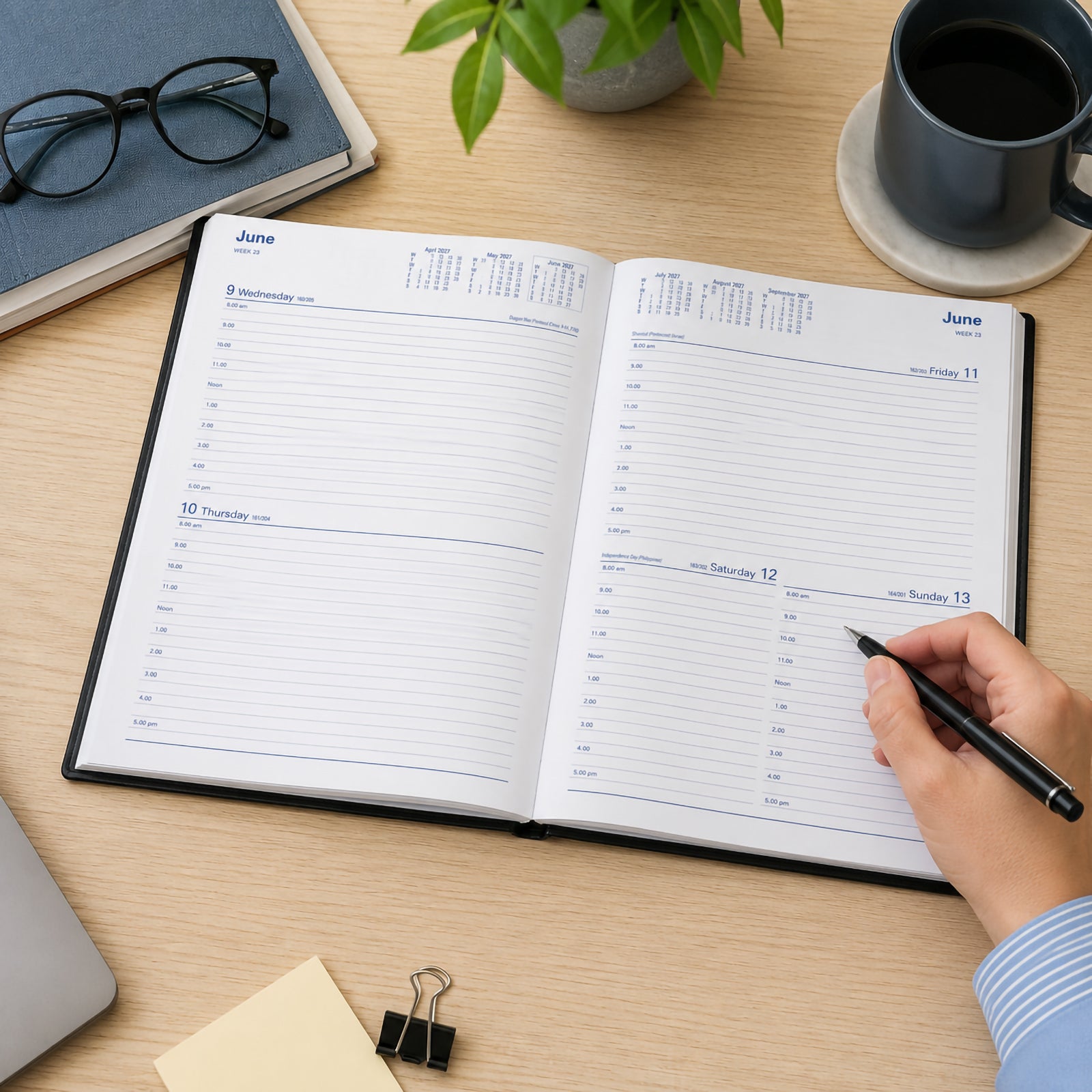 The Collins Belmont 2027 A4 diary is displayed open on a modern wooden office desk while a user writes with a black pen. The spacious two days to a page layout shows hourly scheduling alongside monthly reference calendars. Office accessories including a notebook, coffee mug, glasses and indoor plant create a productive professional workspace.