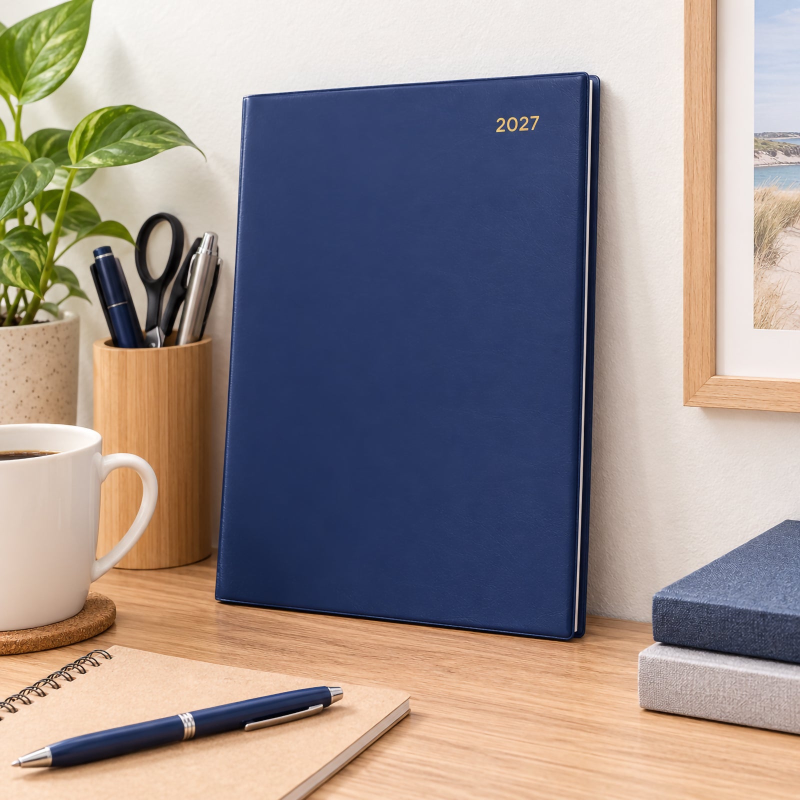 The Collins Belmont 2027 A4 navy diary stands upright on a tidy office desk beside stationery, notebooks, coffee and a leafy plant. The premium navy cover and gold year marking create a refined executive appearance suitable for modern professional workspaces.