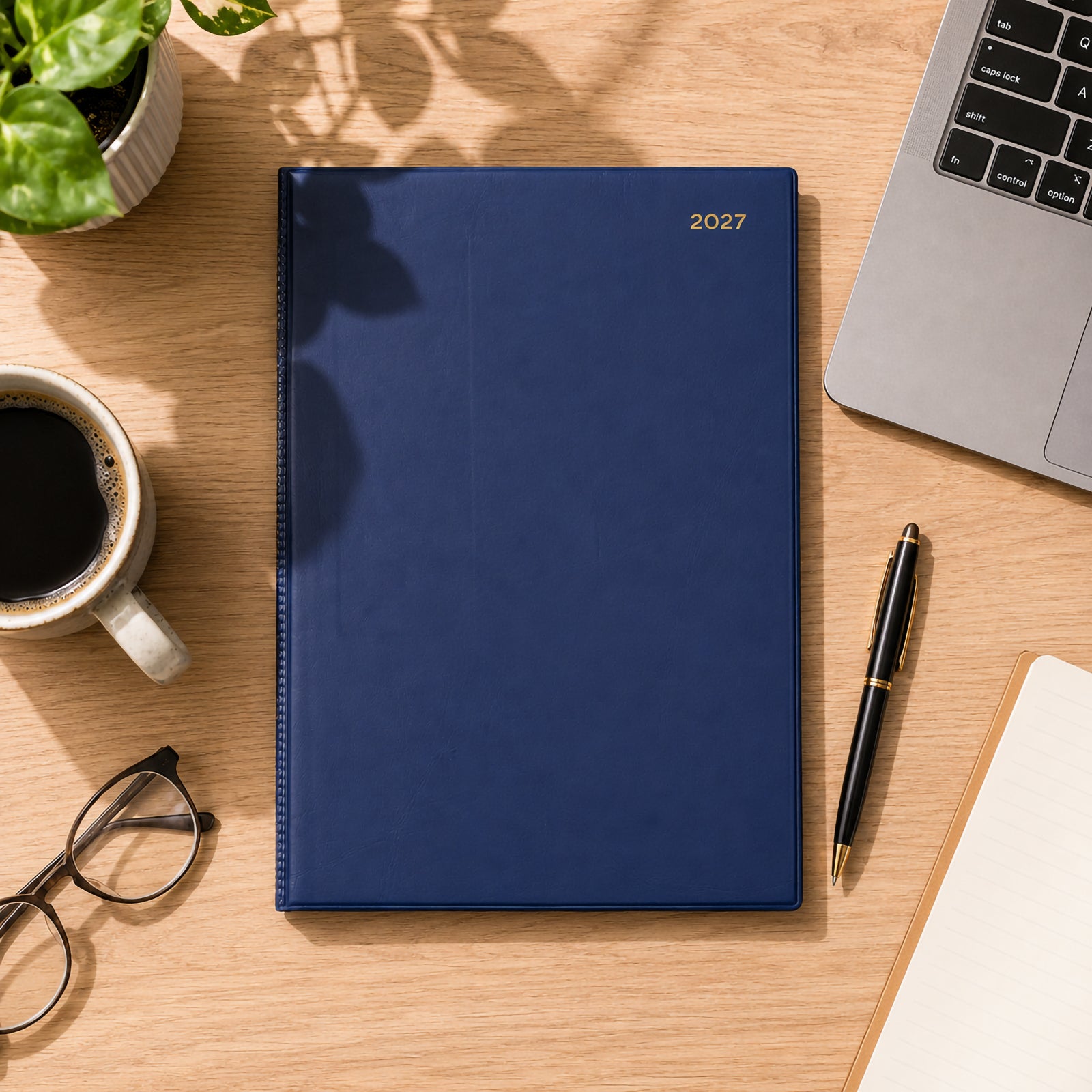 Top view of the Collins Belmont 2027 A4 diary with its premium navy PVC padded cover resting on a timber desk. Natural sunlight, coffee, glasses, laptop, pen and indoor plants create a bright executive workspace that showcases the diary's elegant finish.