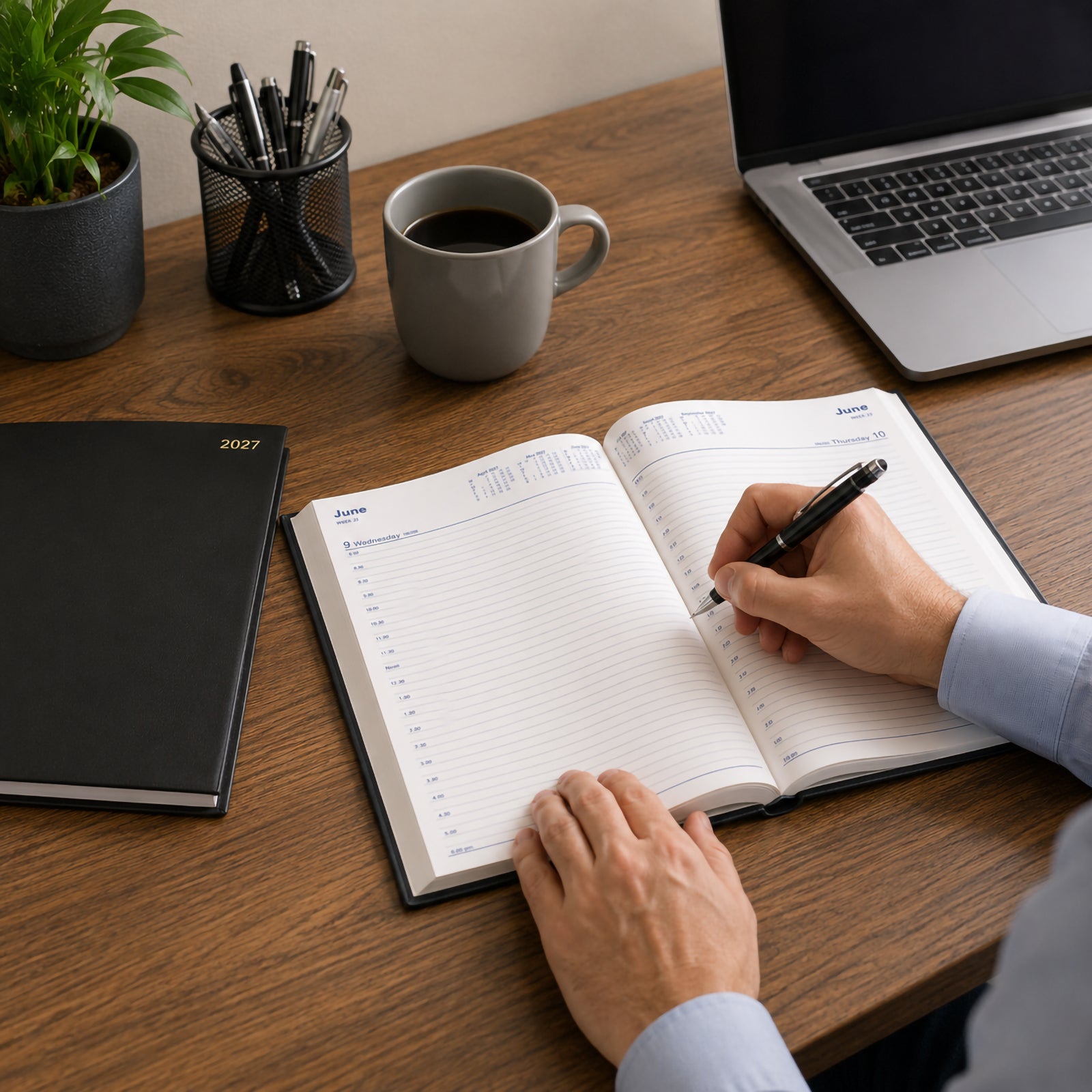 A professional writes in the Collins Belmont 2027 A4 Day to a Page diary at a modern office desk. The spacious daily layout with half hourly appointment scheduling is displayed alongside a laptop, coffee cup, pen holder and office plant, demonstrating productive workplace planning.