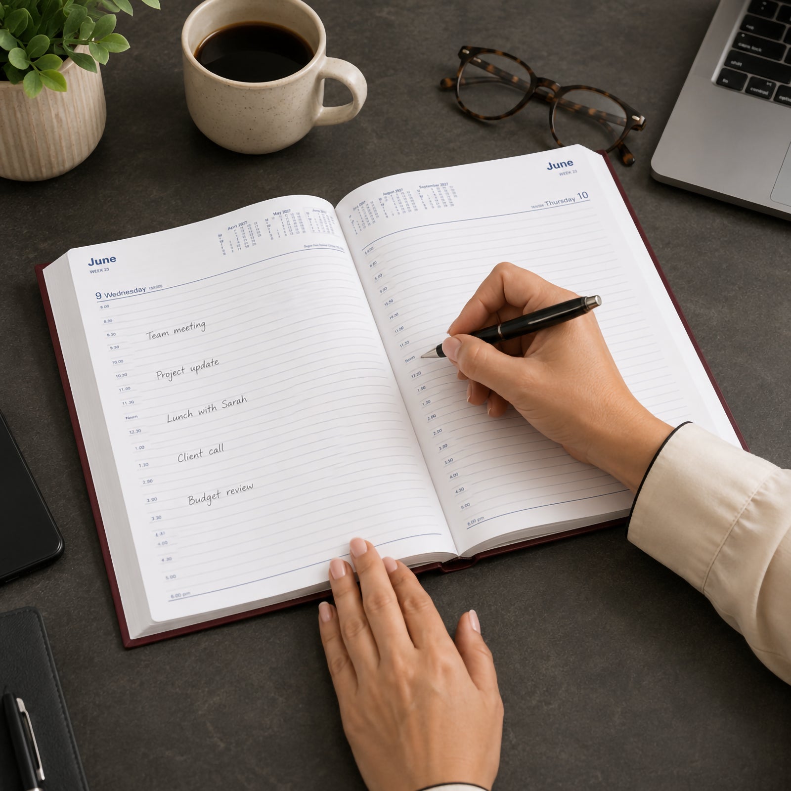 Hands are using the Collins Belmont 2027 A4 Day to a Page Burgundy Diary on a professional workspace, writing appointments into the spacious daily pages. A coffee cup, laptop, glasses and desk accessories create a productive office environment that highlights organised planning.