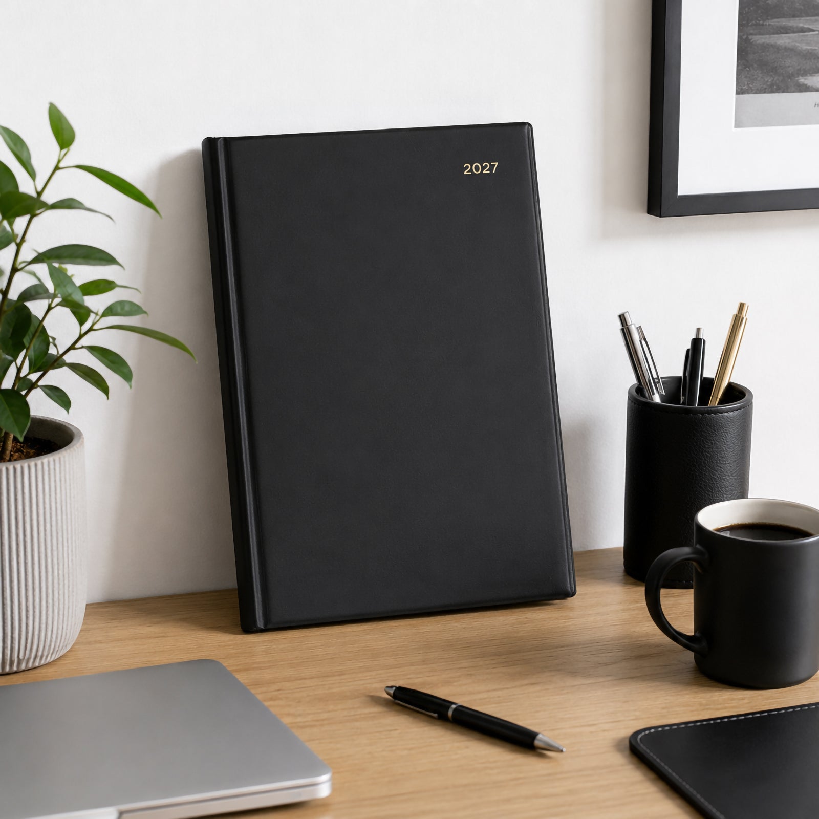 The Collins Belmont 2027 A4 Week to View Black Diary stands upright on an organised office desk beside stationery, a coffee mug, laptop and indoor plant. The executive black cover complements a modern professional office environment.