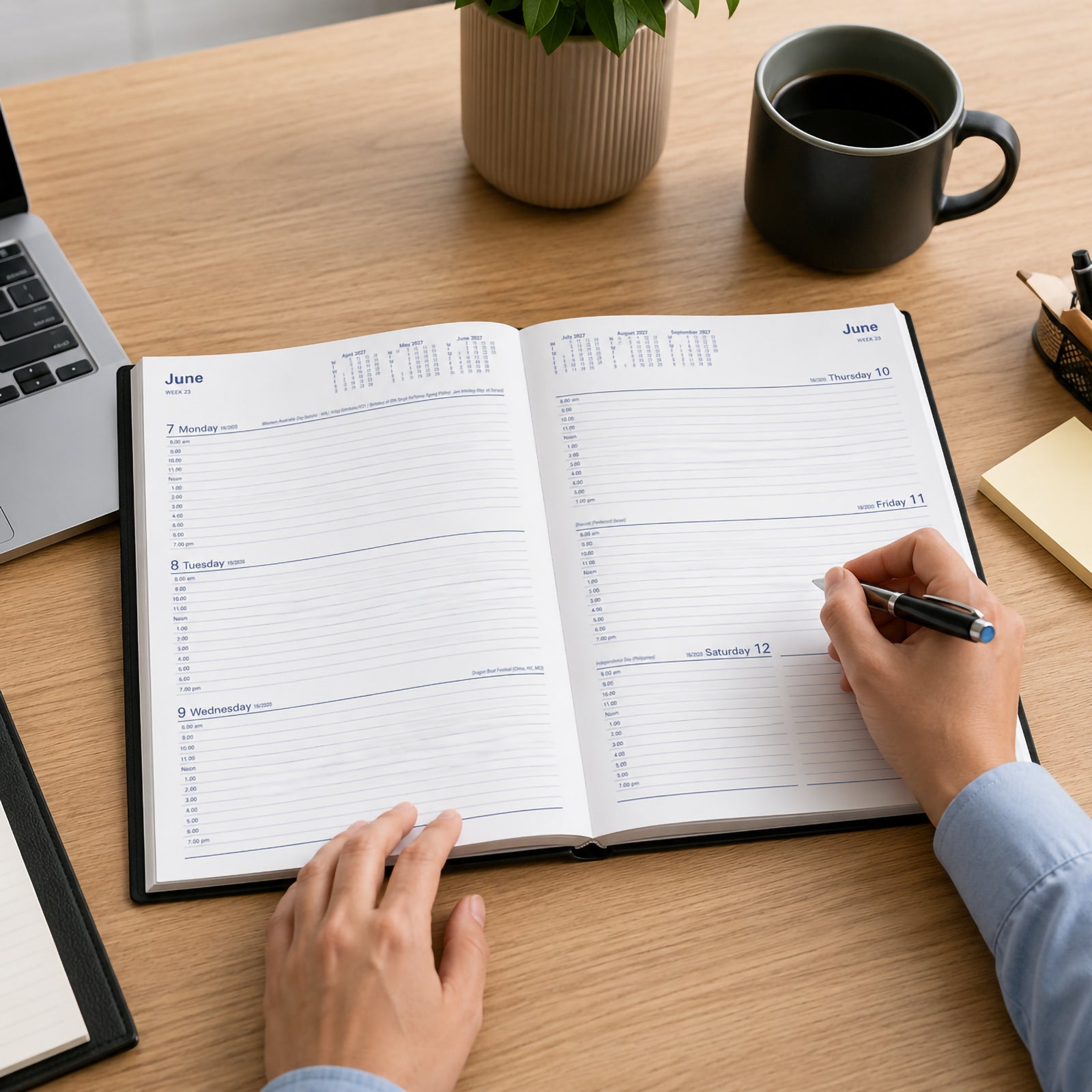 A professional is using the Collins Belmont 2027 A4 Week to View Black Diary on a modern office desk, writing appointments into the spacious weekly planner. The workspace includes a laptop, coffee mug and desk accessories, highlighting productive weekly scheduling.