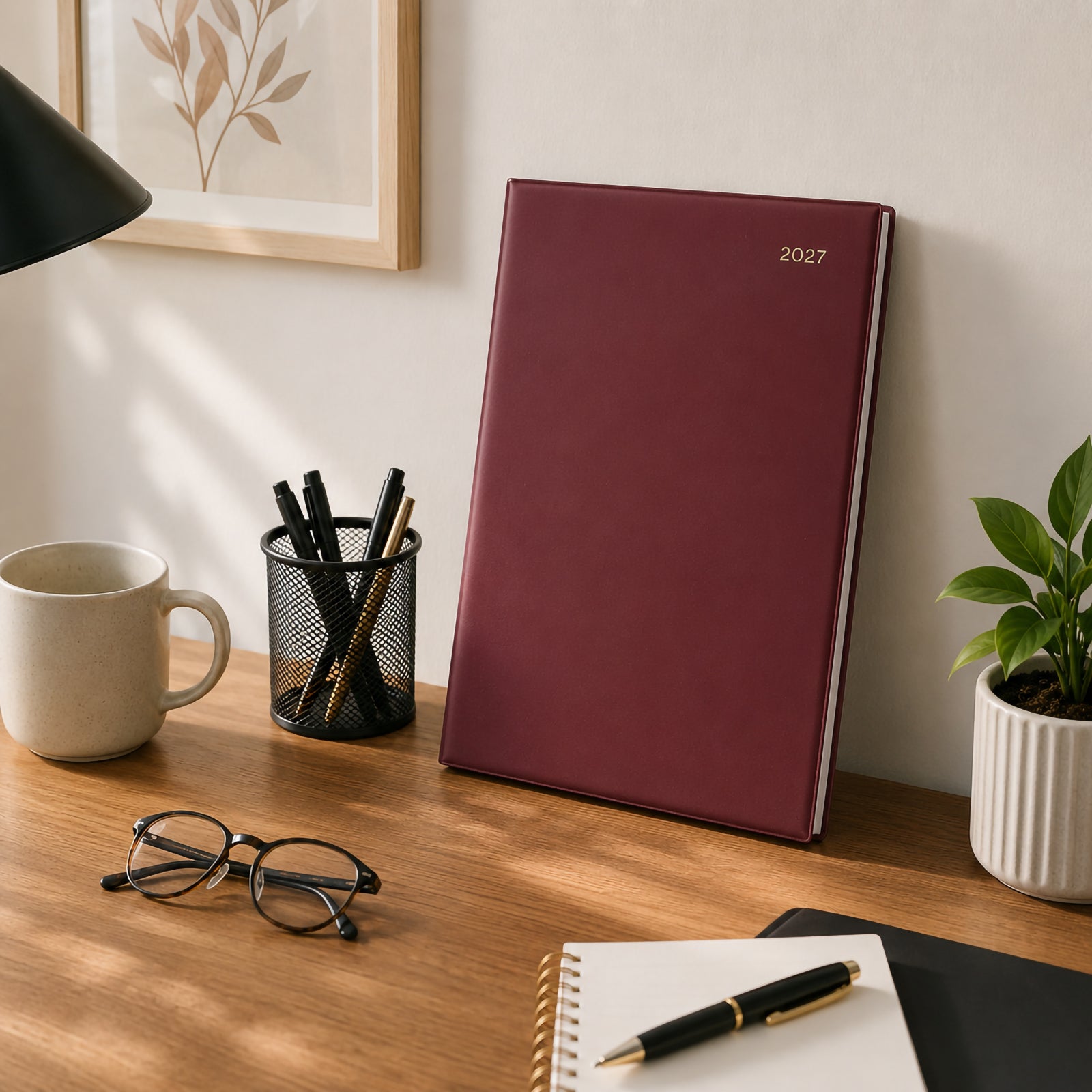 The Collins Belmont 2027 A4 Burgundy Diary stands upright against a wall on a professional office desk surrounded by stationery, indoor plants, a coffee mug and notebook. The elegant burgundy finish complements a sophisticated office environment.