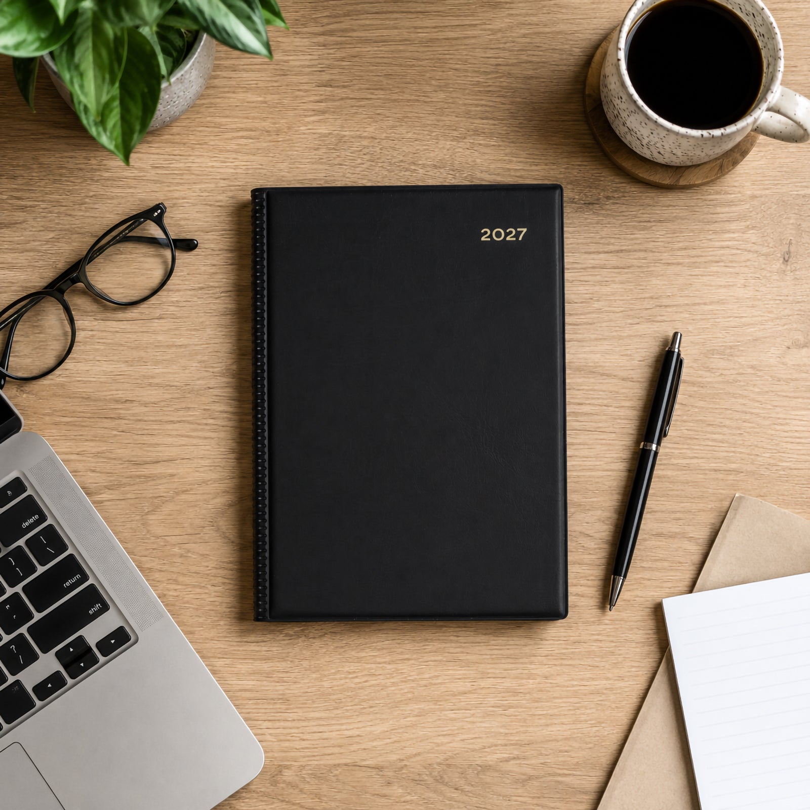 Top view of the Collins Belmont 2027 A5 Black Diary placed neatly on a contemporary office desk beside a laptop, coffee cup, glasses, pen and notebook. The executive black cover complements a clean and organised workspace.