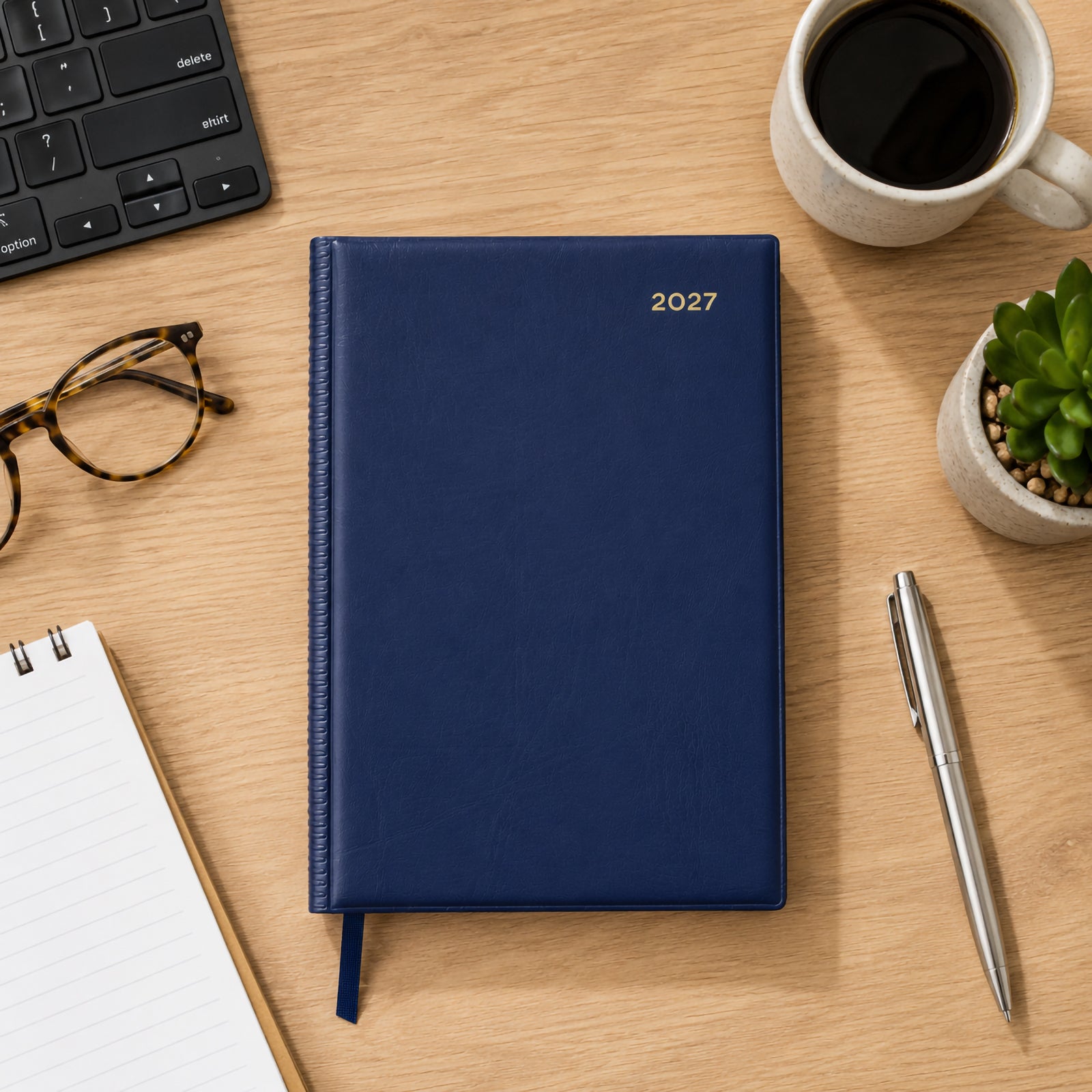 Top view of the Collins Belmont 2027 A5 Navy Diary displayed on a timber office desk beside a notebook, coffee cup, glasses, keyboard, pen and small succulent. The elegant navy cover provides a refined professional appearance.