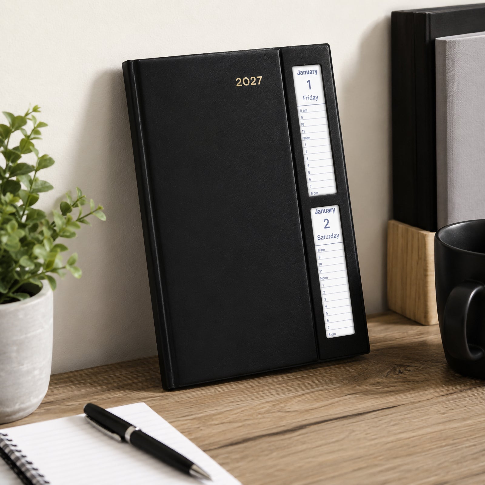 The Collins Belmont 2027 A5 Windowfaced Black Diary stands upright against a wall on a modern office desk with a notebook, coffee mug and indoor plant. The elegant black executive cover and visible date windows create a practical professional presentation.