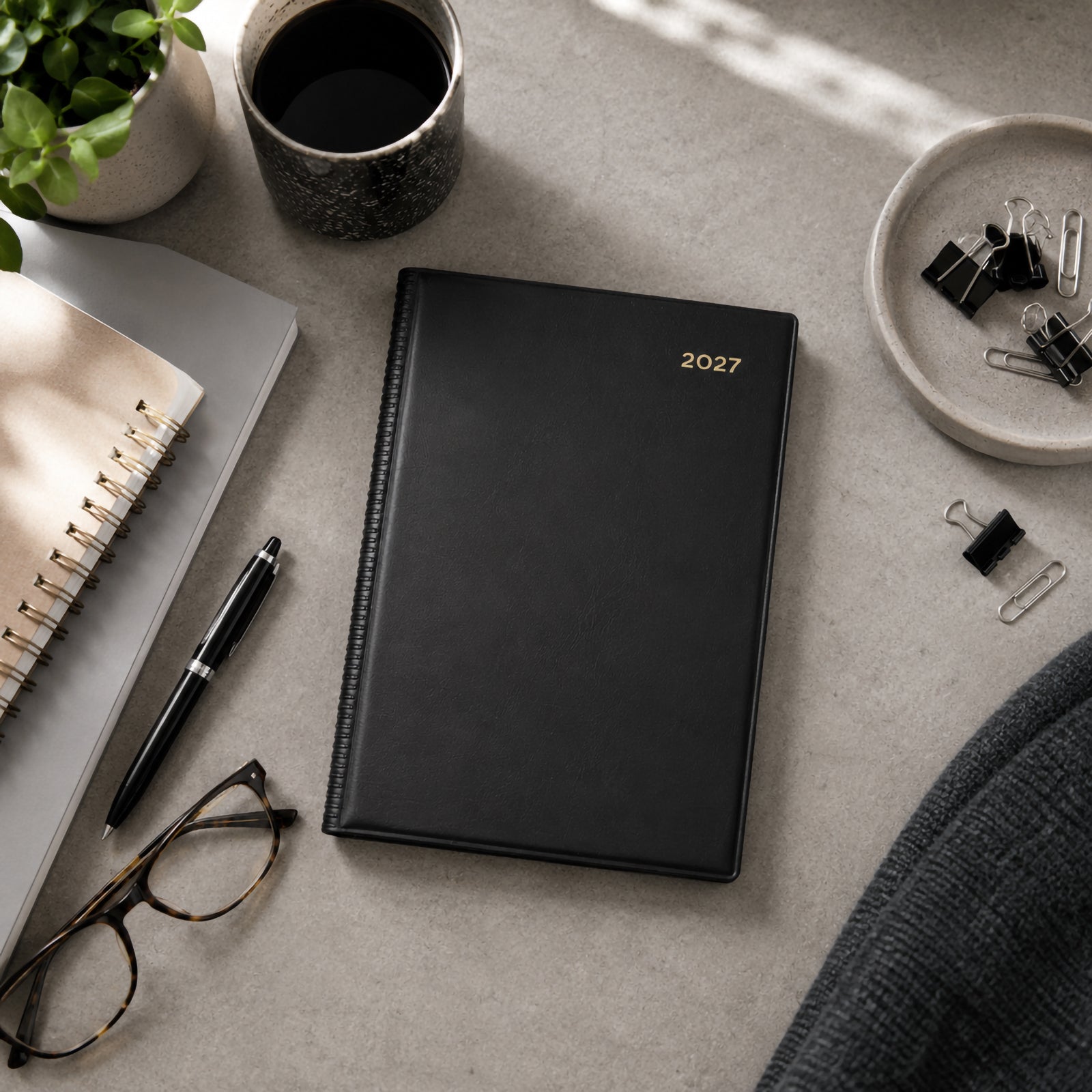 Top view of the Collins Belmont 2027 A5 Day to a Page Black Diary positioned neatly on a contemporary desk with a notebook, coffee cup, pen, glasses and office stationery. The elegant black cover complements a clean and organised professional workspace.