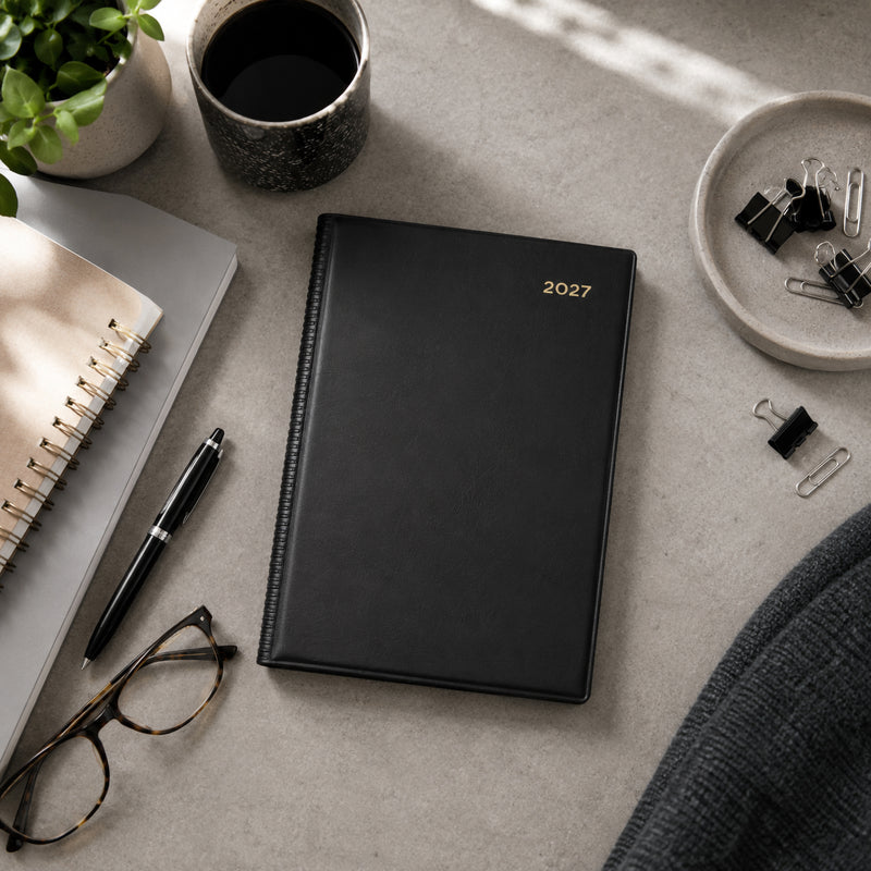 Top view of the Collins Belmont 2027 A5 Day to a Page Black Diary positioned neatly on a contemporary desk with a notebook, coffee cup, pen, glasses and office stationery. The elegant black cover complements a clean and organised professional workspace.