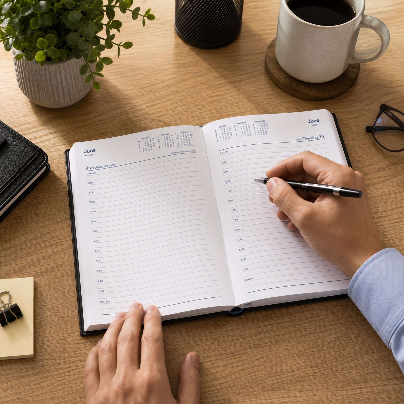 A professional uses the Collins Belmont 2027 A5 Day to a Page Black Diary on a modern timber office desk while writing appointments. A coffee mug, notebook, glasses and desk accessories create a productive workspace that demonstrates the diary in everyday business use.