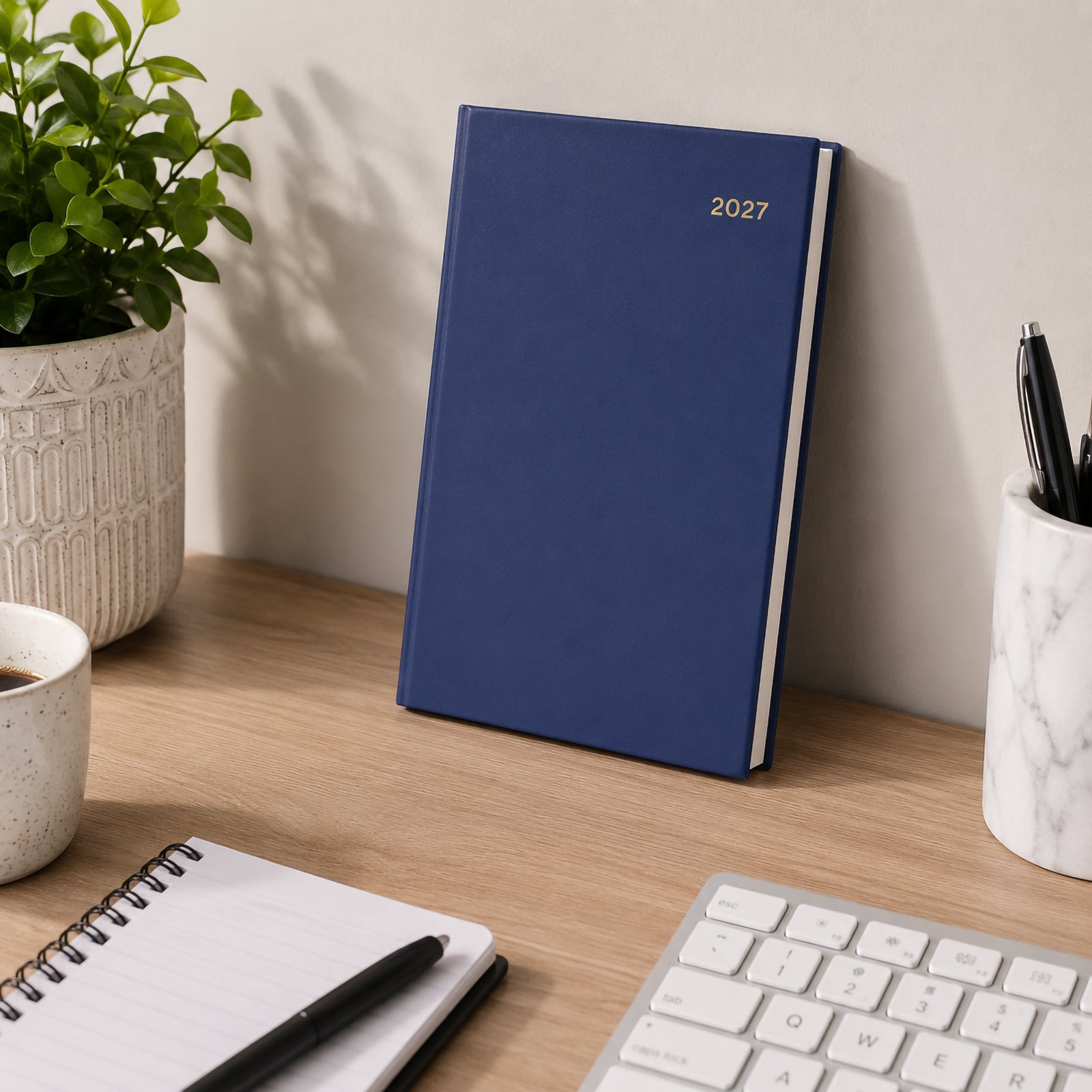 The Collins Belmont 2027 A5 Day to a Page Navy Diary standing upright against a wall on a modern office desk with stationery, keyboard, notebook, coffee and decorative greenery. The lifestyle scene showcases its premium appearance in a professional environment.