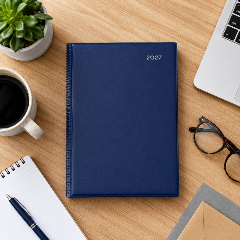 Top view of the Collins Belmont 2027 A5 Day to a Page Navy Diary positioned on a wooden desk beside a laptop, coffee cup, notebook, pen and reading glasses. The image highlights the professional navy cover in a clean and organised workspace.