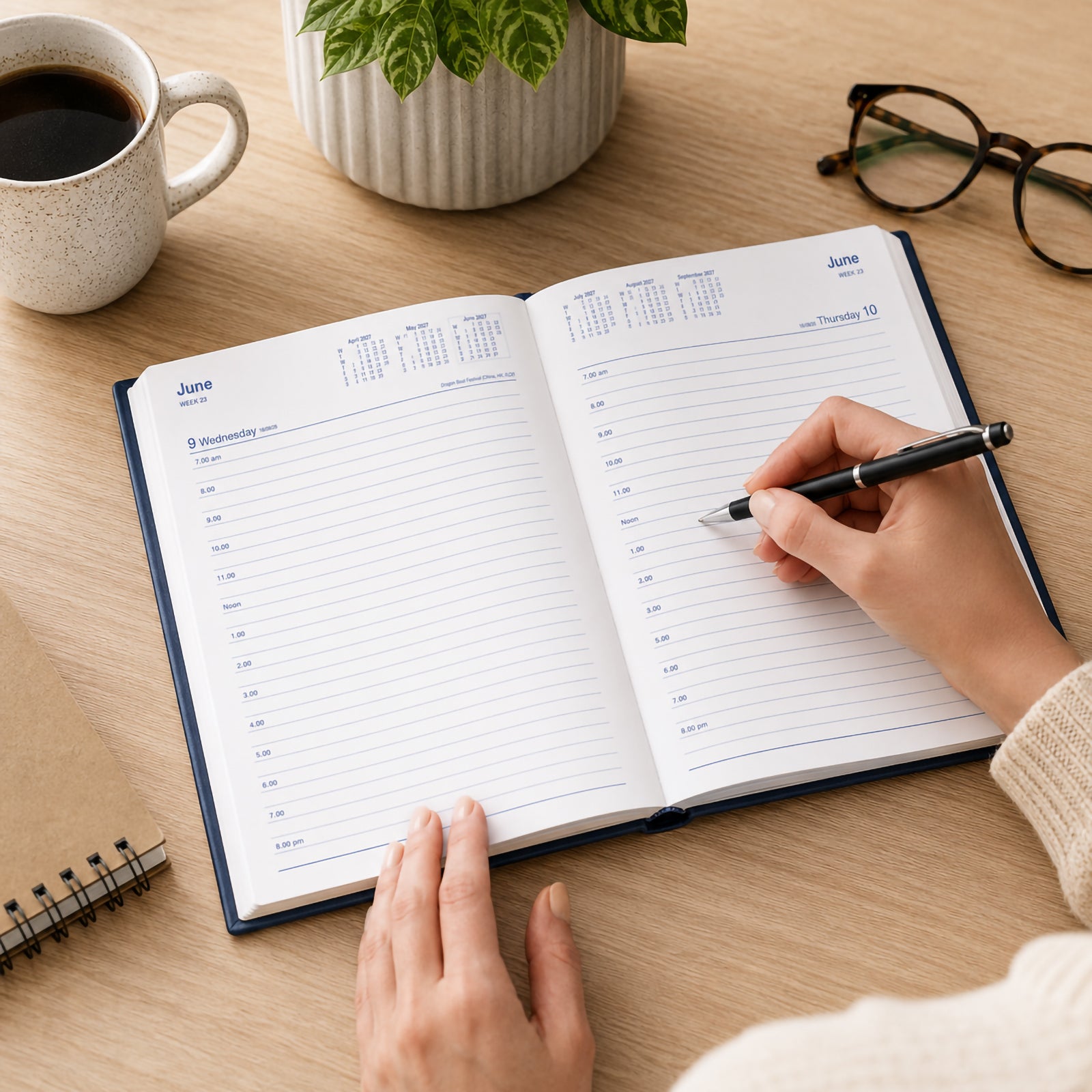 A person writes in the Collins Belmont 2027 A5 Day to a Page Navy Diary at a tidy timber desk surrounded by coffee, a notebook, reading glasses and indoor plants. The spacious daily layout demonstrates how the diary supports organised appointments, schedules and professional planning.