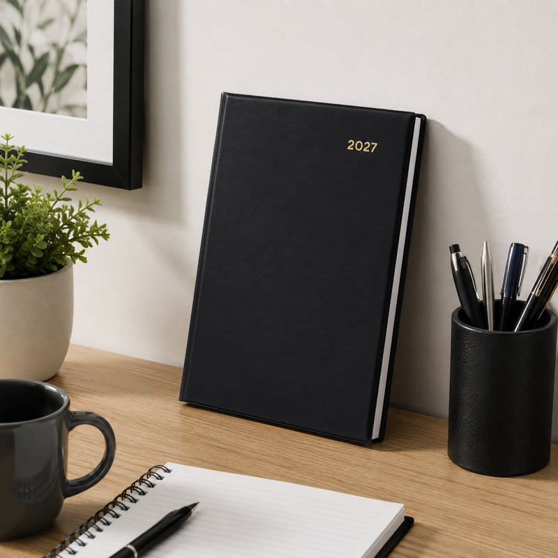 The Collins Belmont 2027 A5 Week to View Black Diary displayed upright against a wall on a stylish office desk with notebook, coffee mug, pen holder and indoor plant. The elegant black finish highlights its premium professional appearance.
