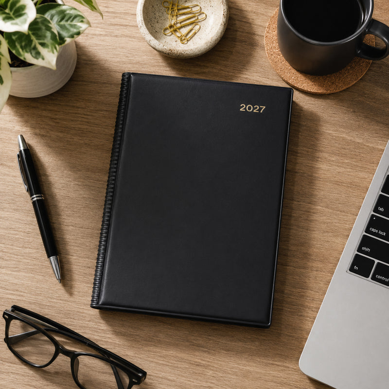 Top view of the Collins Belmont 2027 A5 Week to View Black Diary positioned on a timber desk beside a laptop, coffee cup, stationery, decorative plant and reading glasses. The professional black cover complements modern office and home workspaces.