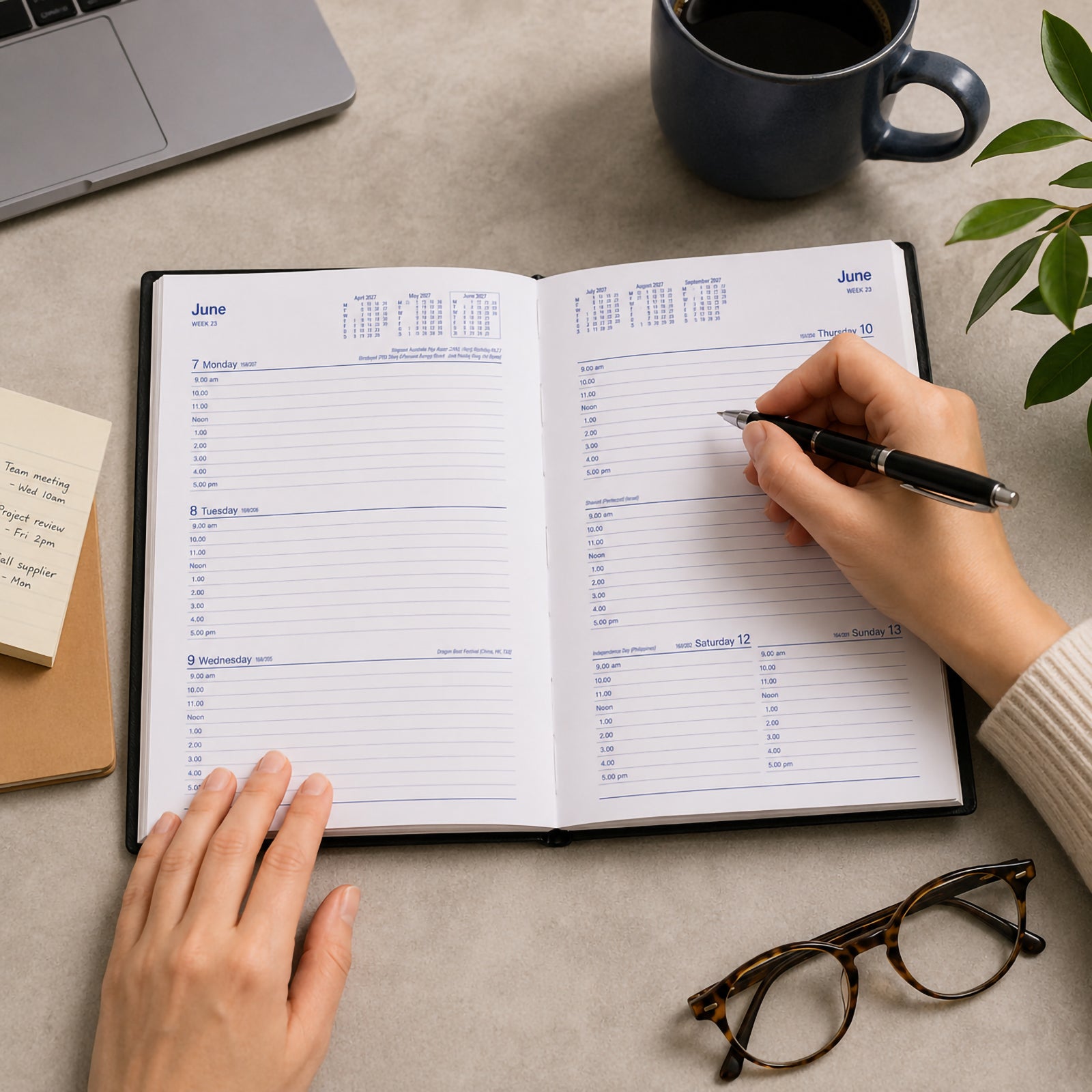 A person writes in the Collins Belmont 2027 A5 Week to View Black Diary on a contemporary workspace featuring a laptop, coffee mug, notebook and glasses. The weekly layout provides a clear overview of appointments, making it ideal for business planning and organised daily productivity.