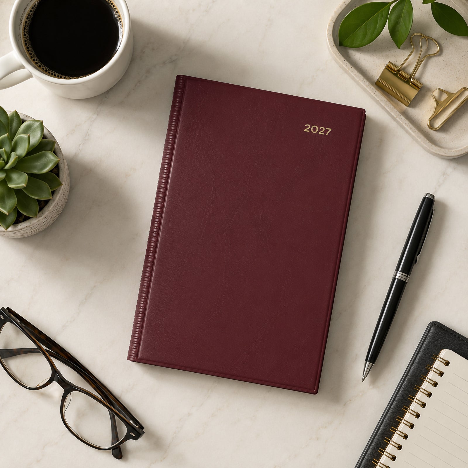 Top view of the Collins Belmont 2027 A5 Week to View Burgundy Diary displayed on a light marble desk with coffee, stationery, succulent, glasses and notebook. The elegant burgundy cover creates a refined and professional workspace presentation.