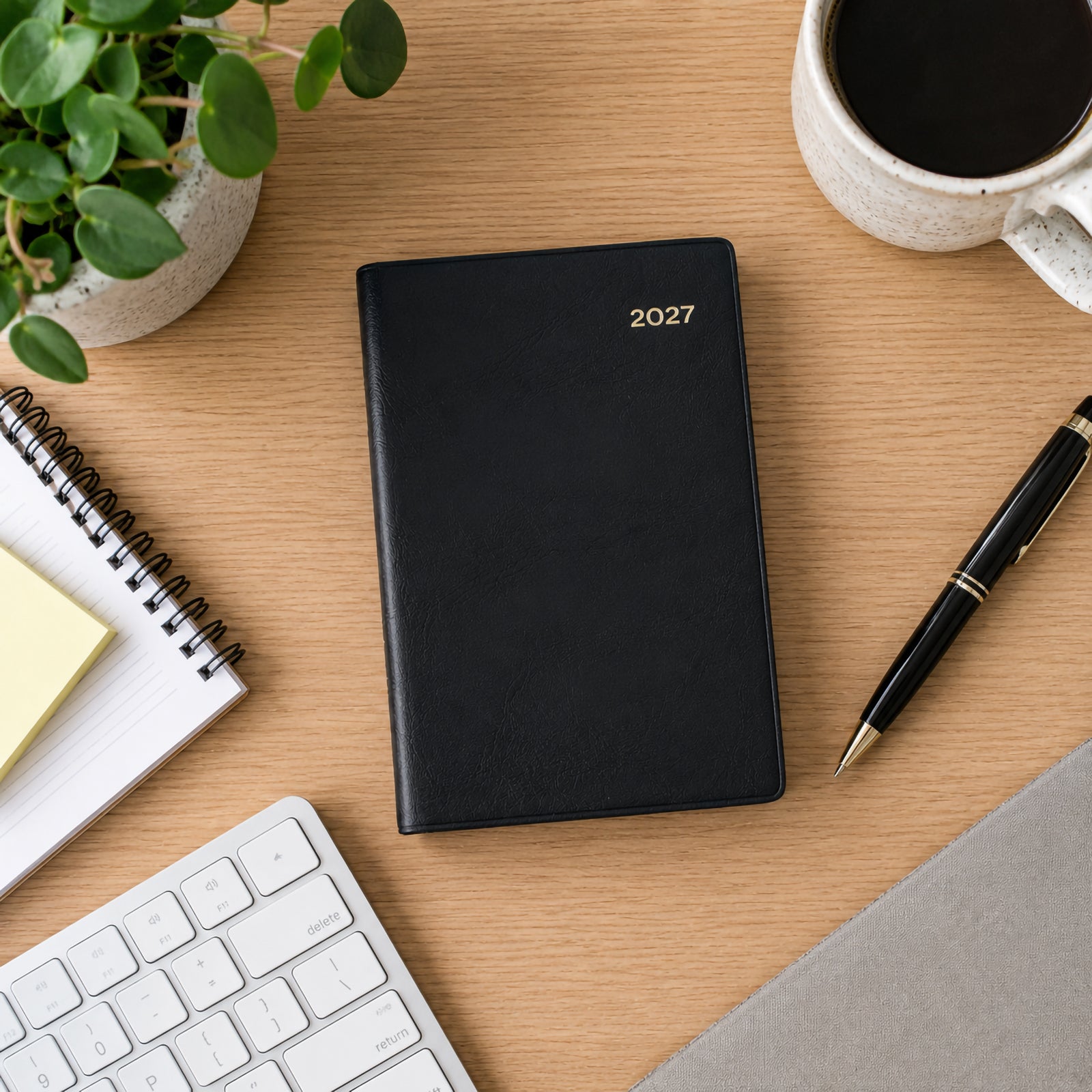 Top view of the Collins Belmont 2027 A7 Pocket Diary with its black PVC cover displayed on a modern timber desk beside a keyboard, coffee mug, notebook, pen and indoor plant. The compact professional design is ideal for organised daily planning.