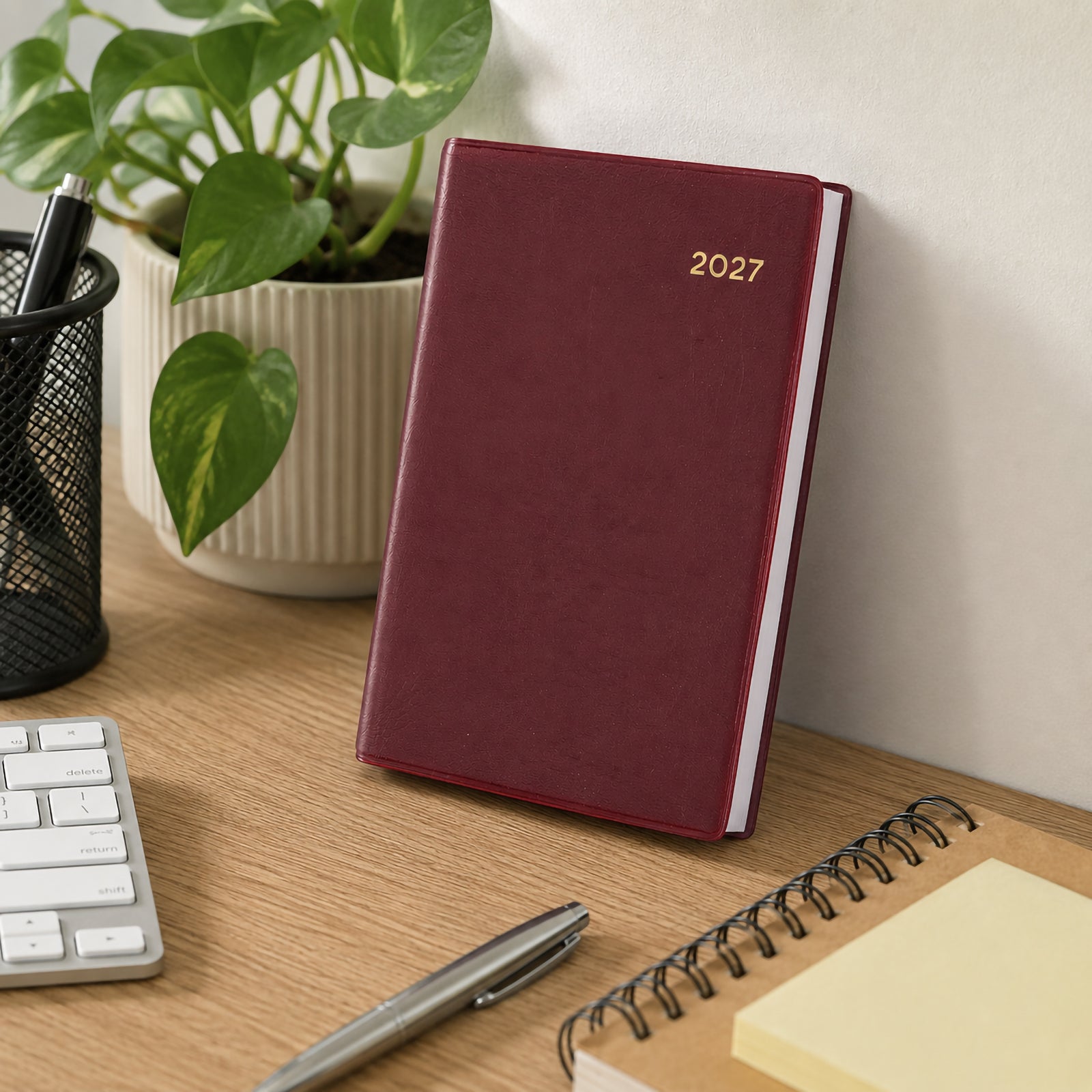 The Collins Belmont 2027 A7 Pocket Diary in burgundy stands upright against a wall on a tidy office desk surrounded by a keyboard, notebook, pen holder and decorative plant. The premium burgundy cover complements a modern workspace while emphasising durability and compact convenience.