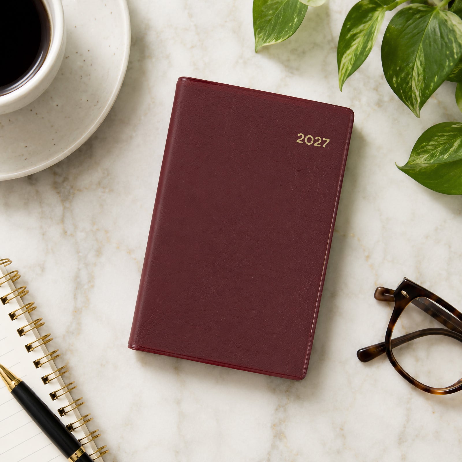 Top view of the Collins Belmont 2027 A7 Pocket Diary with its elegant burgundy PVC cover placed on a marble desk beside a coffee cup, notebook, glasses and indoor plant. The refined arrangement showcases the diary as a stylish organiser for everyday planning and professional use.