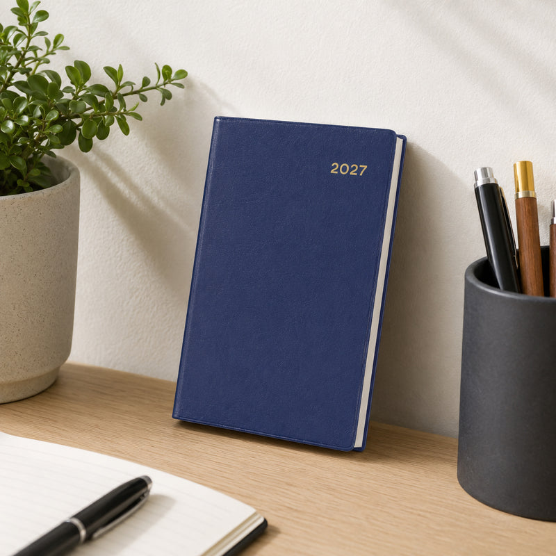 The Collins Belmont 2027 A7 Pocket Diary in navy stands upright against a wall on a tidy office desk beside a pen holder, notebook and decorative plant. The sophisticated navy cover complements a modern workspace while emphasising portability and professional style.