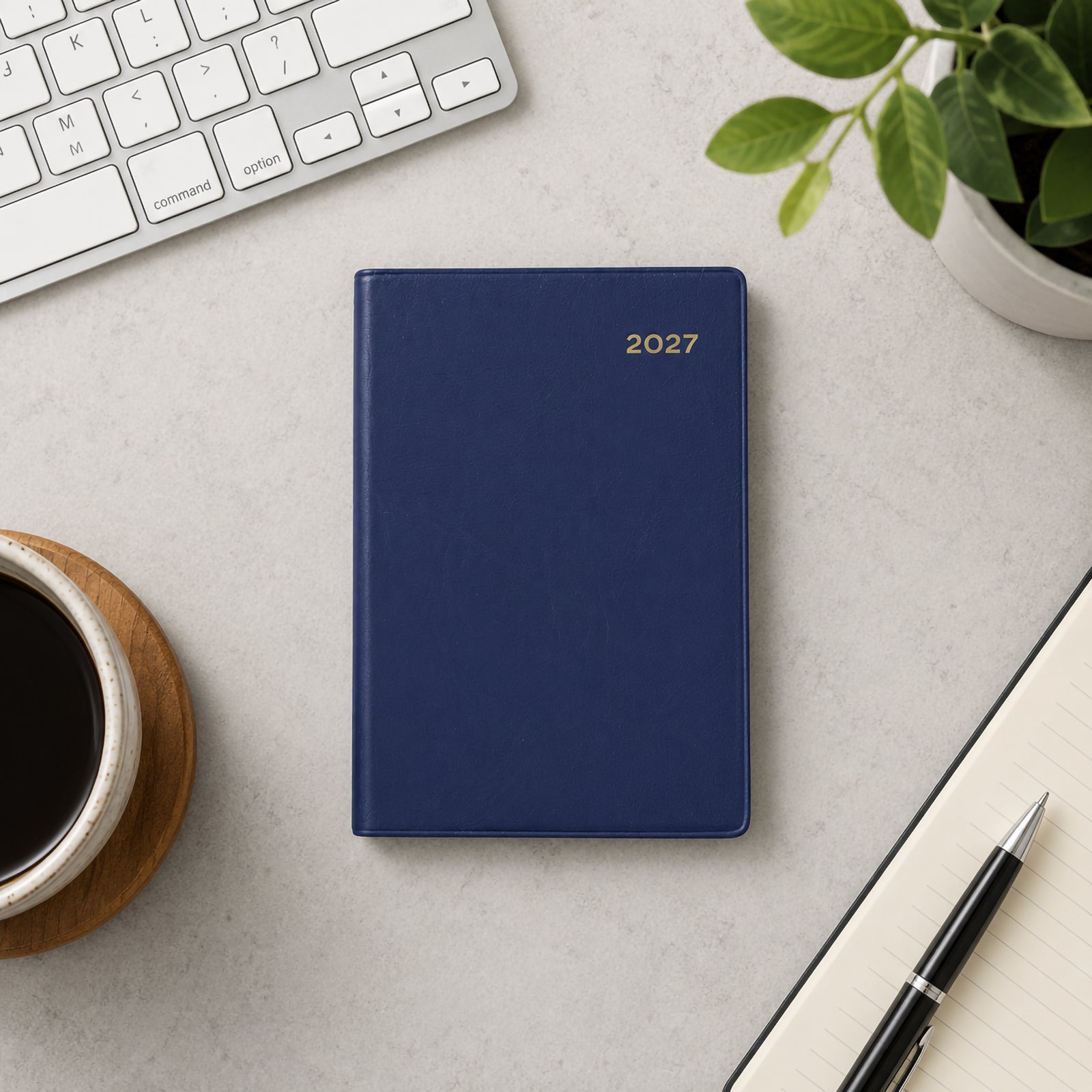 Top view of the Collins Belmont 2027 A7 Pocket Diary with its elegant navy PVC cover displayed beside a keyboard, coffee cup, notebook, pen and indoor plant. The neat workspace showcases the diary as a practical organiser for professional and personal planning.
