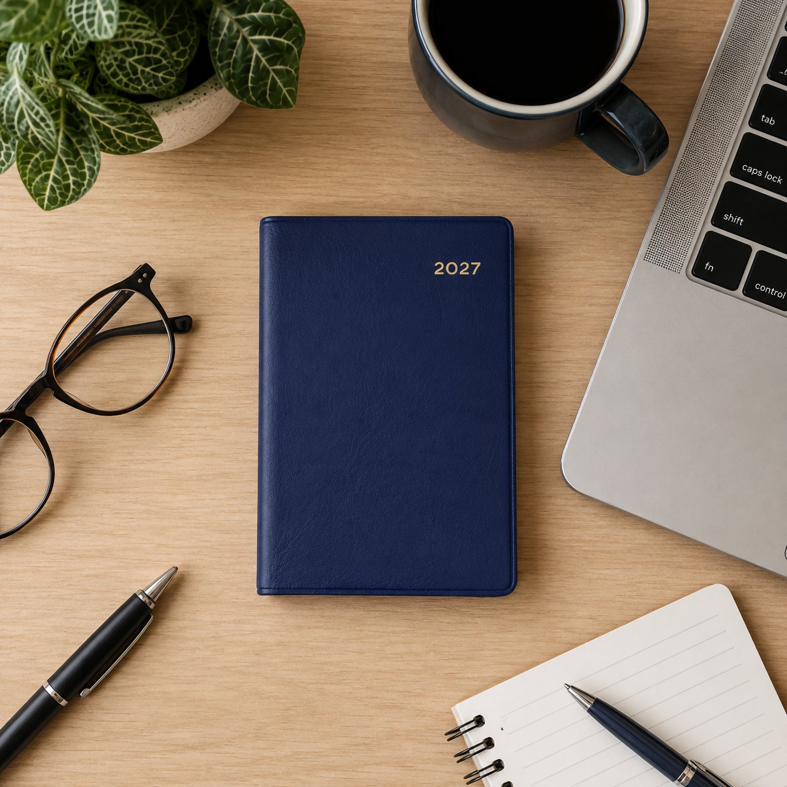 Top view of the Collins Belmont 2027 A7 Week to View Pocket Diary in navy displayed on a timber desk beside a laptop, coffee cup, glasses, notebook, pen and indoor plant. The workspace highlights portable organisation and professional style.