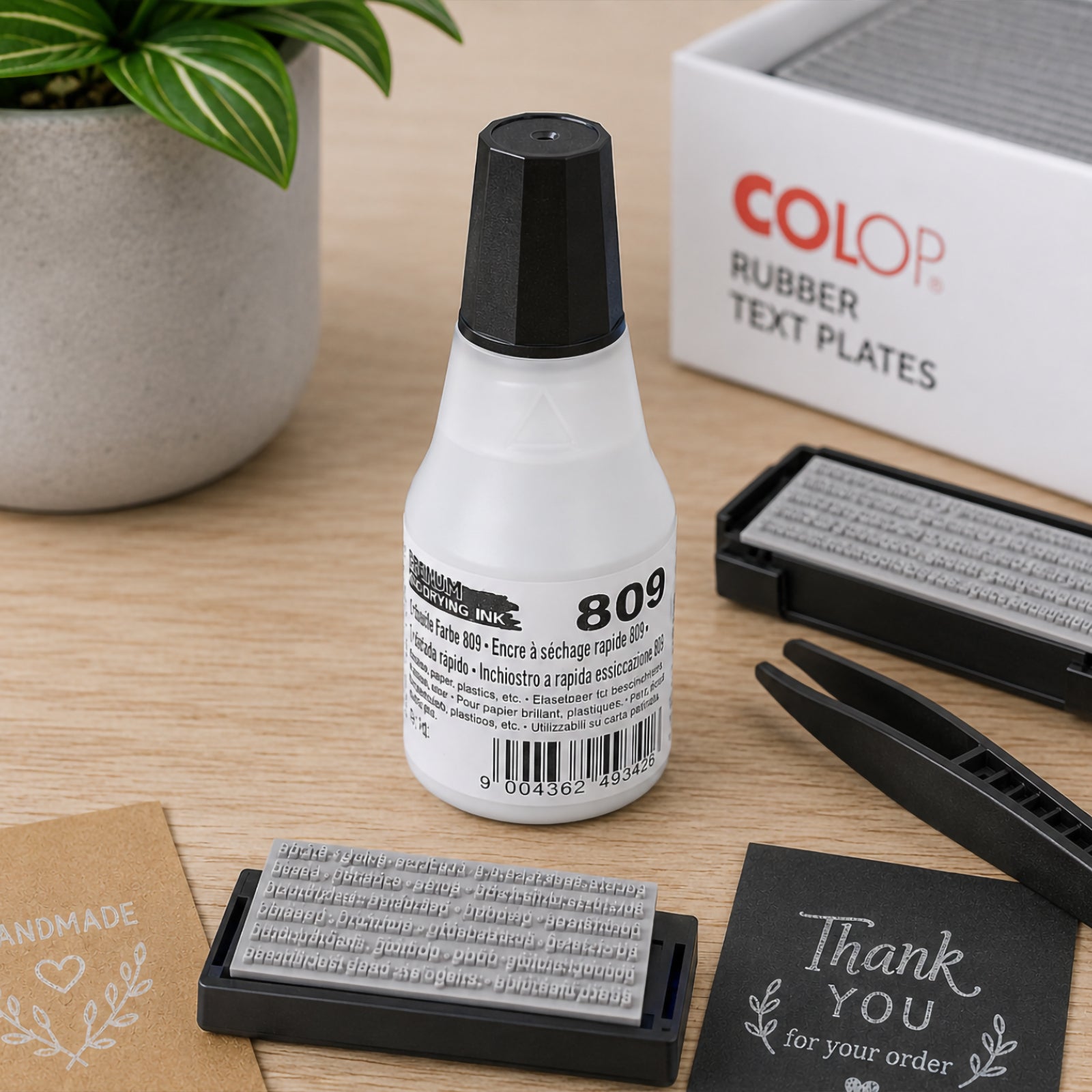 Colop 809 Quick Drying Ink Premium White 25ml bottle displayed on an office desk beside rubber text plates, tweezers and handmade packaging materials, highlighting its suitability for custom stamp creation and precision refilling.
