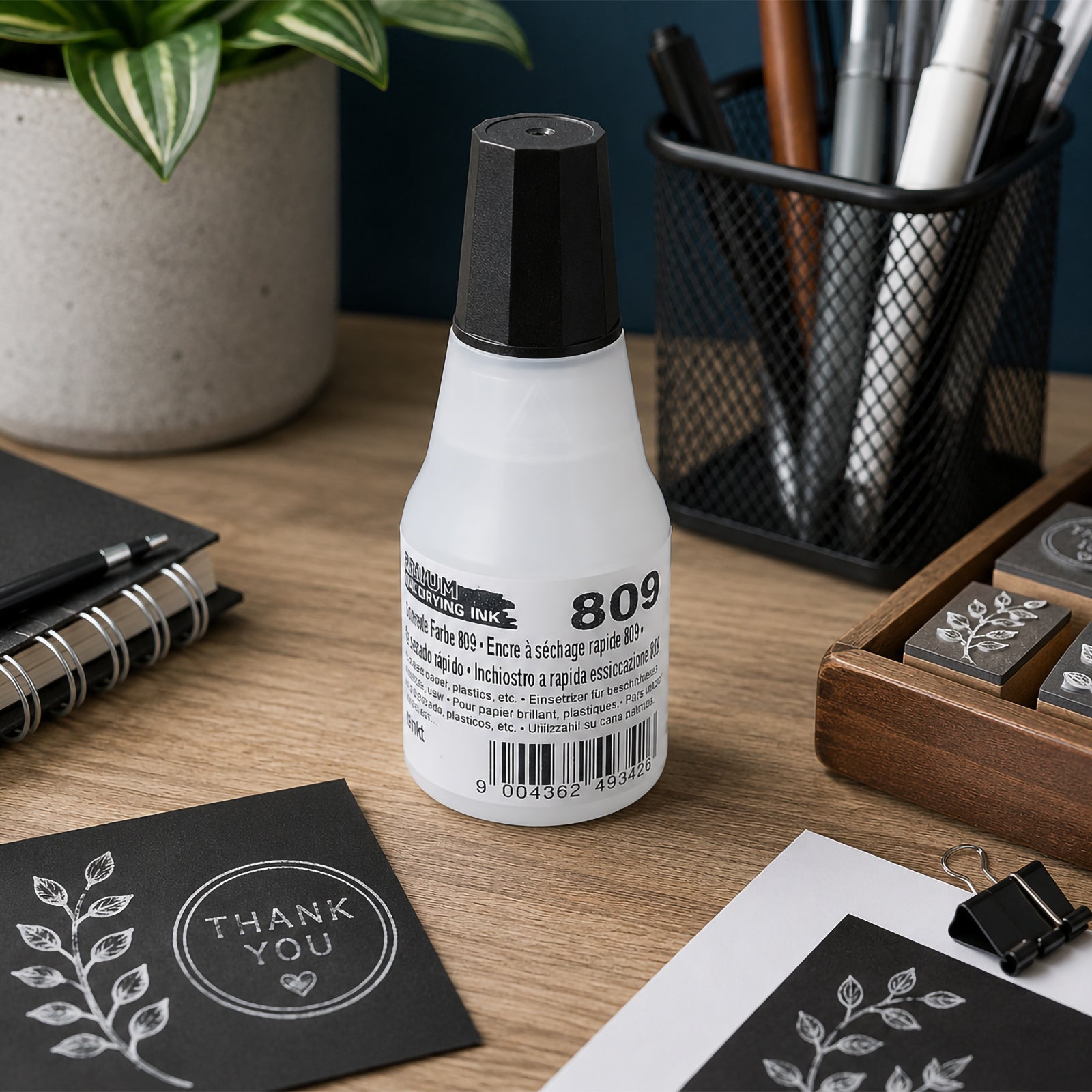 Lifestyle image of the Colop 809 Quick Drying Ink Premium White 25ml bottle positioned on a desktop beside notebooks, rubber stamps and stationery, illustrating everyday use in office, creative studio and craft environments.