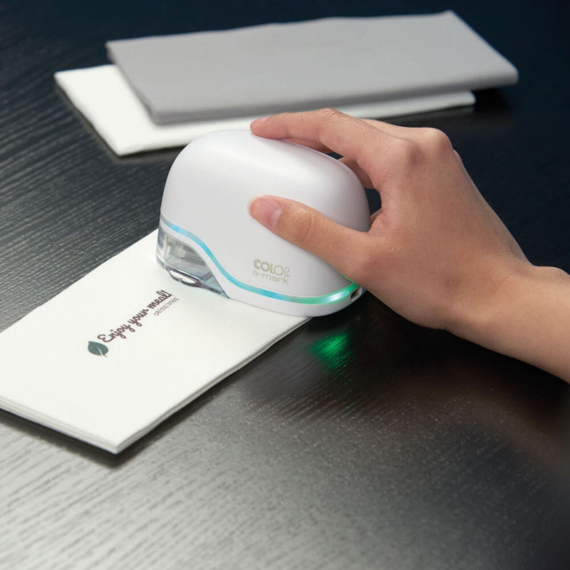 Hand sweeping the white Colop e-mark across a folded white napkin, printing a small logo and message in one pass