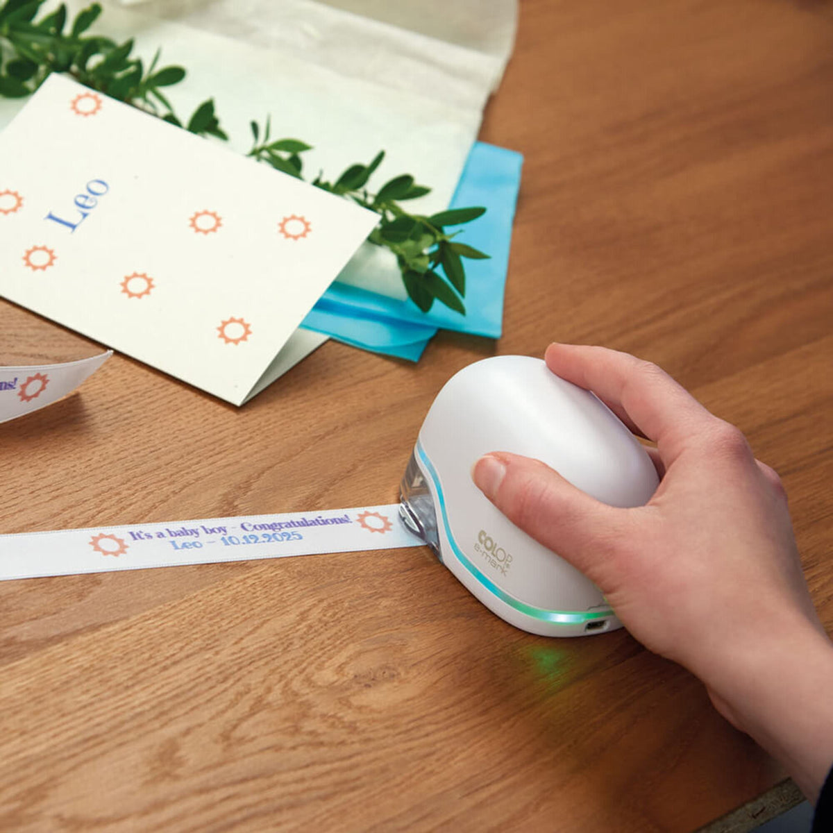 Hand printing a name ribbon with the white Colop e-mark on a wooden table beside printed gift cards and envelopes