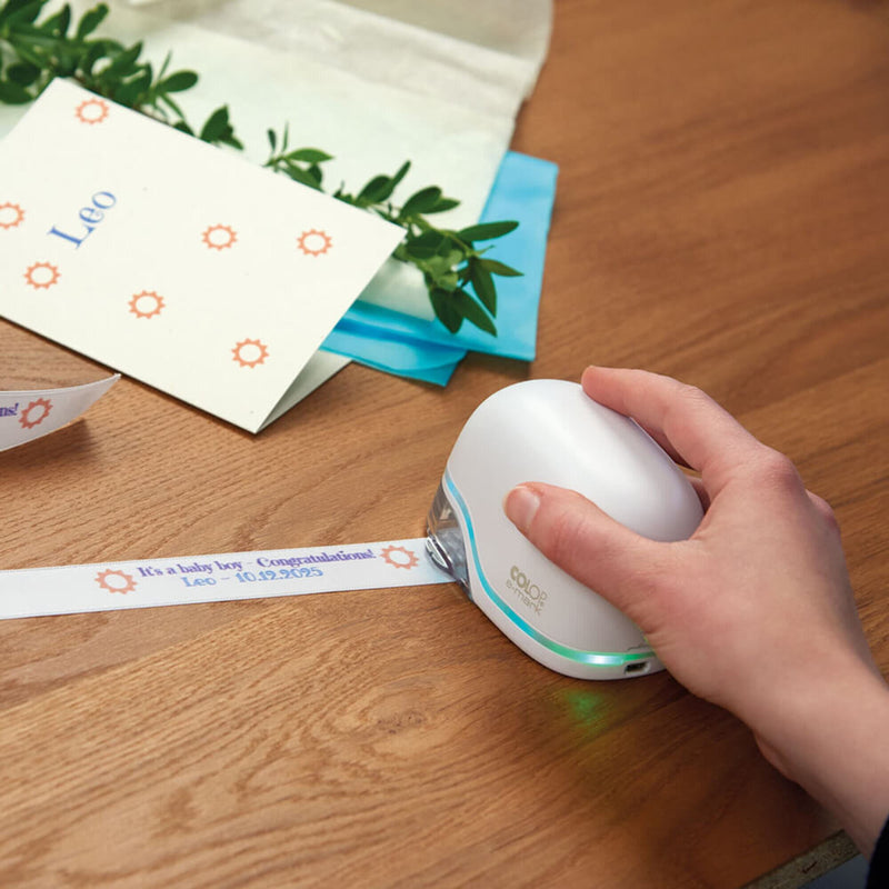 Hand printing a name ribbon with the white Colop e-mark on a wooden table beside printed gift cards and envelopes