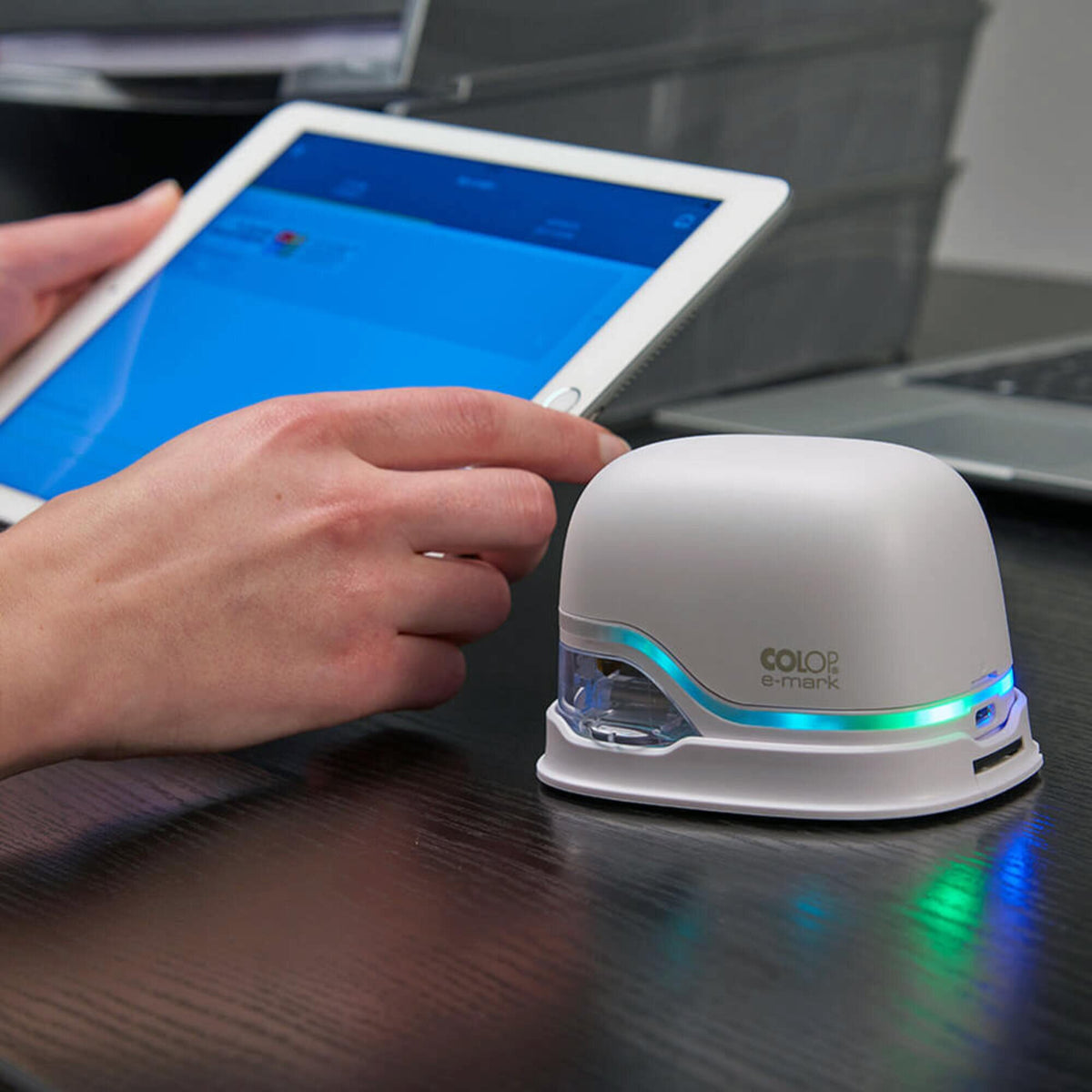 Finger tapping a tablet running the Colop app while the white e-mark handheld printer sits beside it on a dark desk