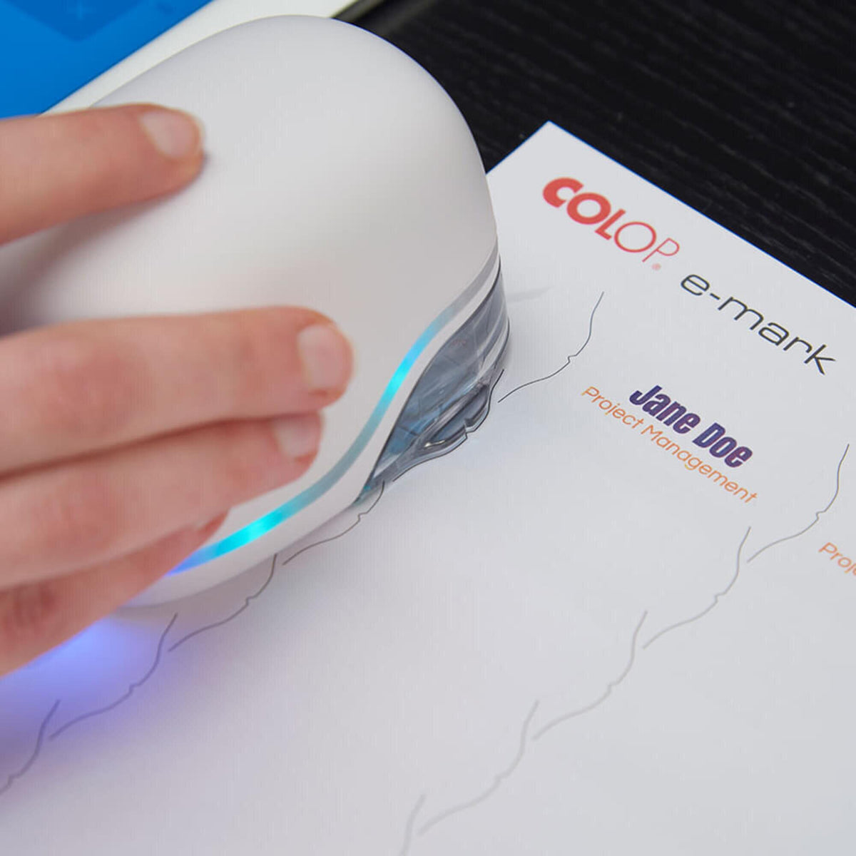 Close view of a hand sweeping the white Colop e-mark across paper, printing a coloured Colop e-mark logo and a name