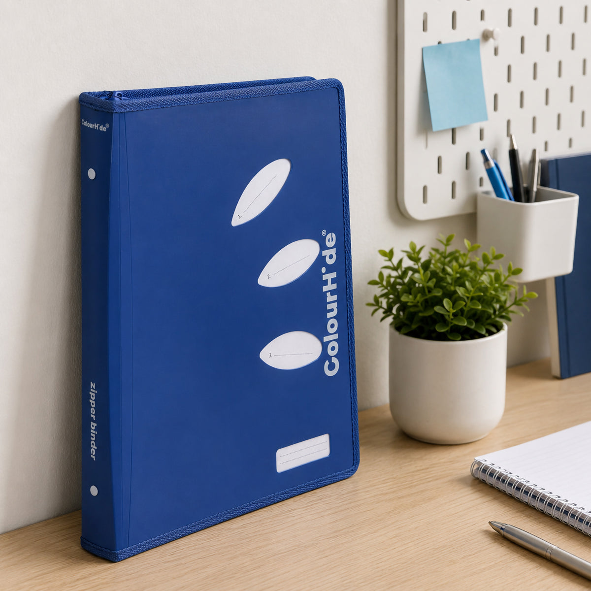 Illustrative desk scene from our earlier listing: a blue zipper binder leaning against a pegboard wall beside a pot plant