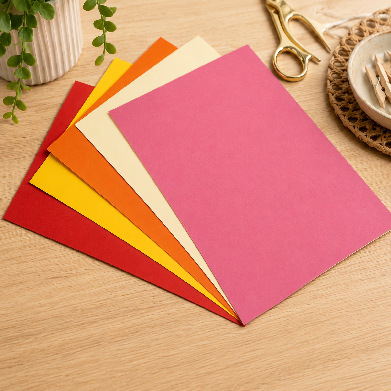 Selection of assorted warm coloured board sheets arranged in a fan pattern on a wooden desk. The visible colours include red, yellow, orange, cream and pink, showcasing the variety of tones available for classroom displays, seasonal decorations and craft projects.