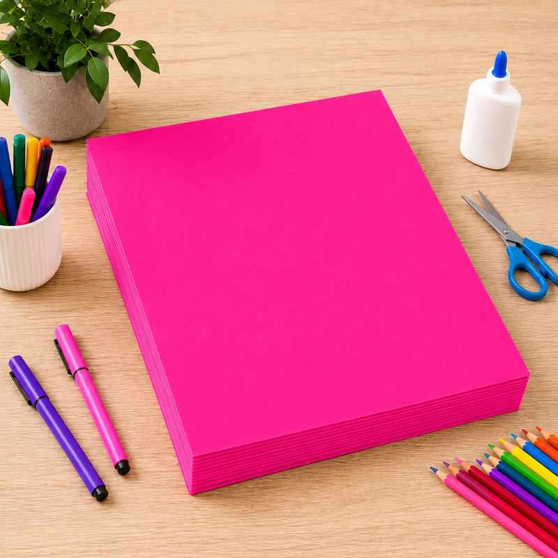Neatly stacked sheets of fluro pink 250gsm project board displayed on a wooden craft table alongside markers, coloured pencils, scissors and glue. The image highlights the board's vibrant fluorescent colour, sturdy construction and suitability for posters, displays and creative educational projects.