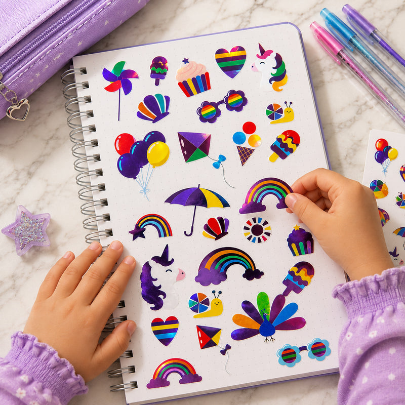 Open sketchbook decorated with the full Colours of Joy sticker collection. Vibrant rainbows, peacocks, kites, hearts and unicorn designs are arranged across the page, showcasing the bright colours and reflective foil finish that make every project stand out.