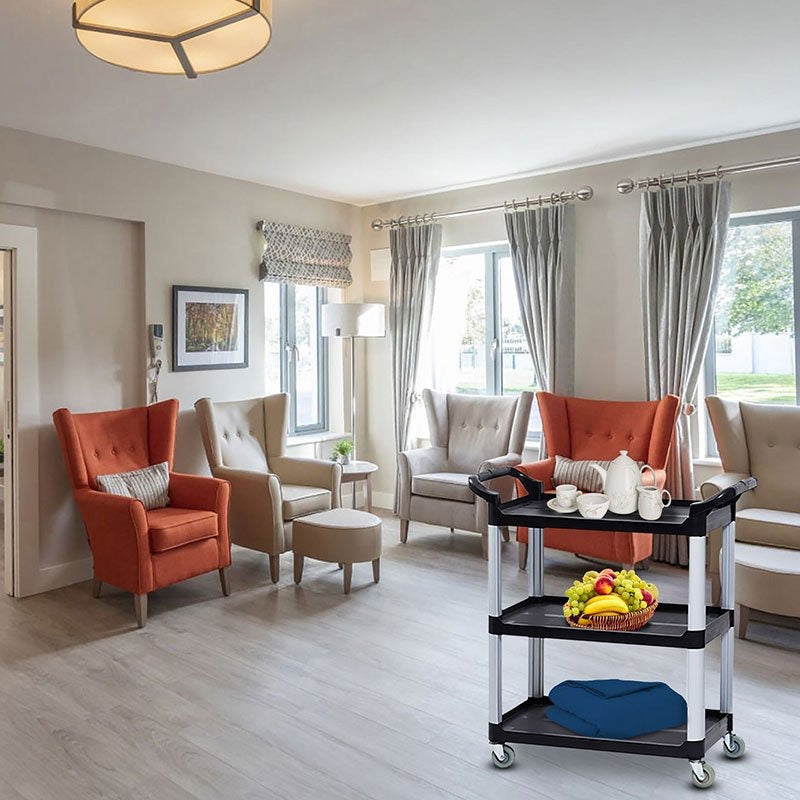 The Compass 7224941 compact utility cart in an aged care lounge room, carrying a tea set on the top shelf and a fruit basket on the middle shelf, beside upholstered armchairs.