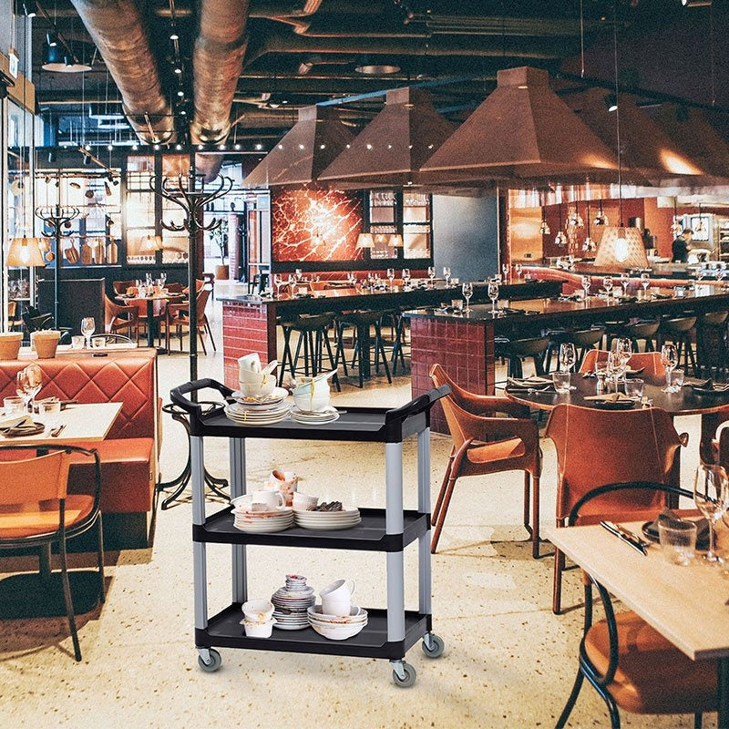 The Compass 7224941 compact utility cart in a restaurant interior, loaded with stacked crockery on its shelves beside timber tables and bar seating.
